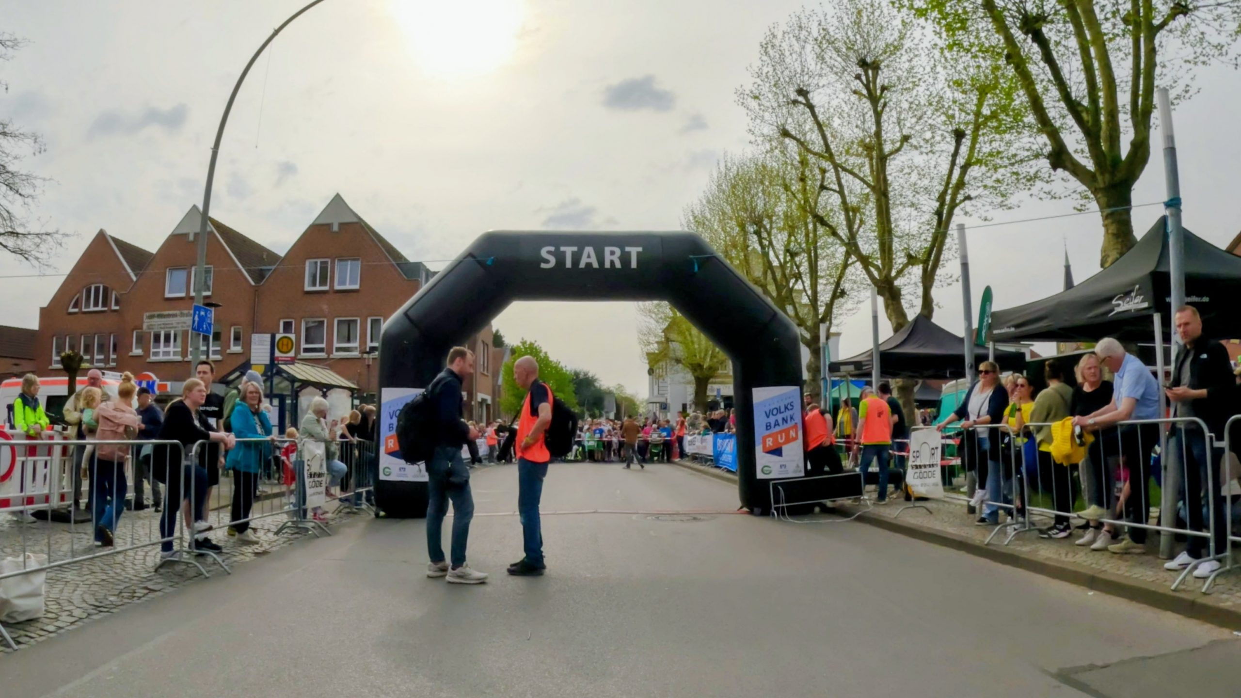 Two runners chat at the start line under a black inflatable arch marked START, as spectators stand behind barriers along a town street.