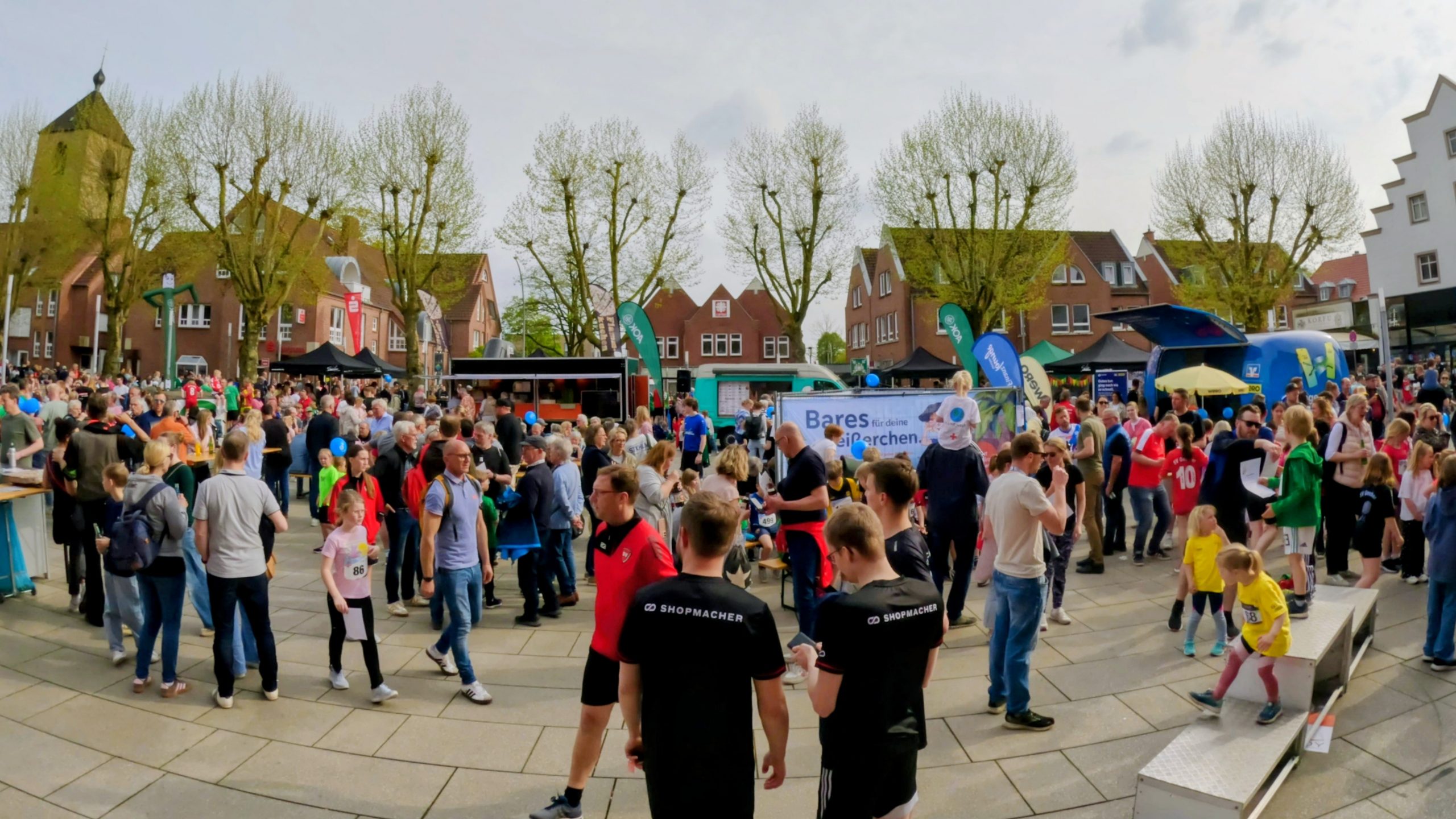 Crowd of people at an outdoor festival in a town square with tents, banners, and trees in the background.
