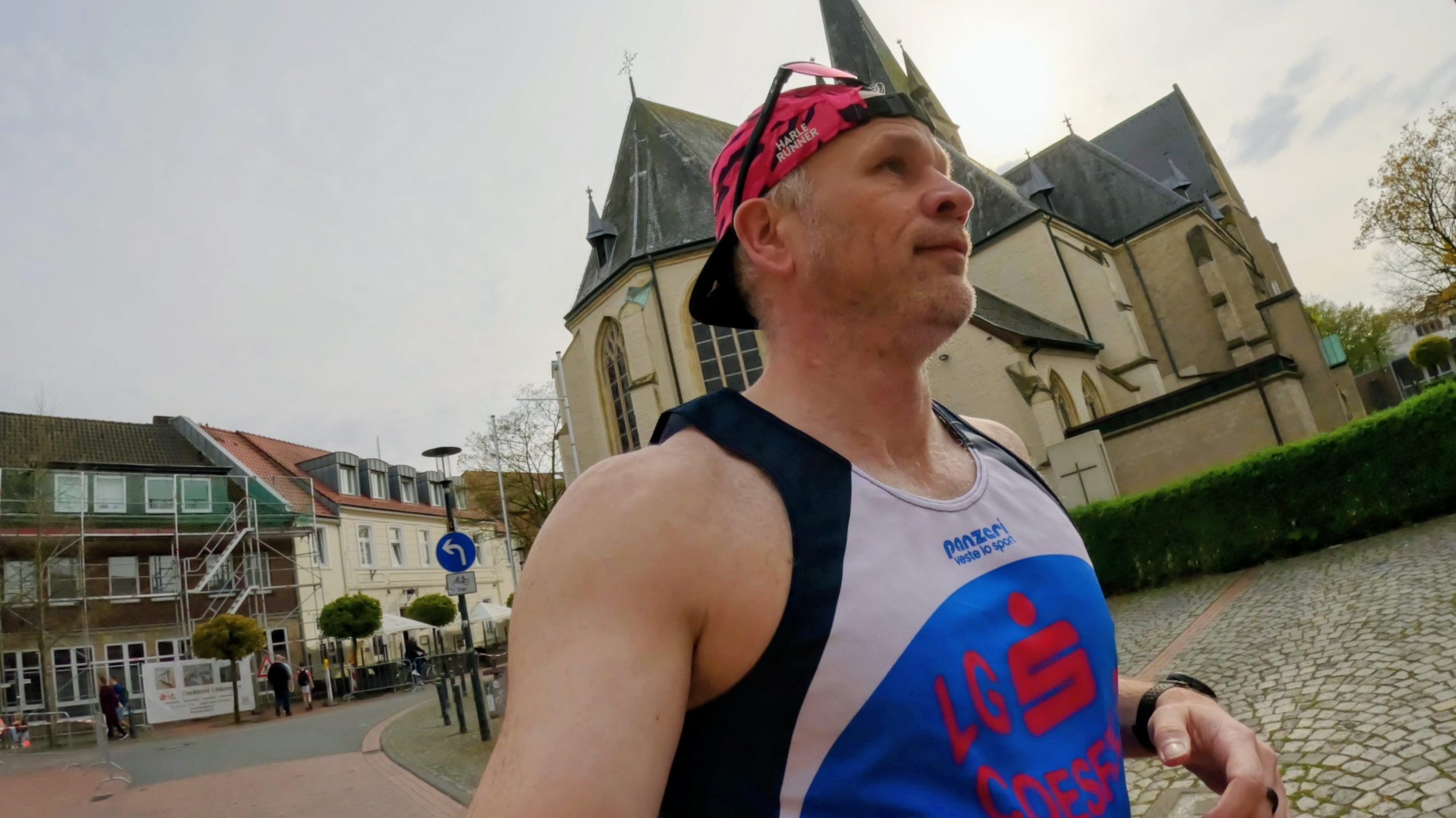 Male runner in a pink headband and blue-white singlet runs along a cobblestone street with a church in the background in an urban race setting.