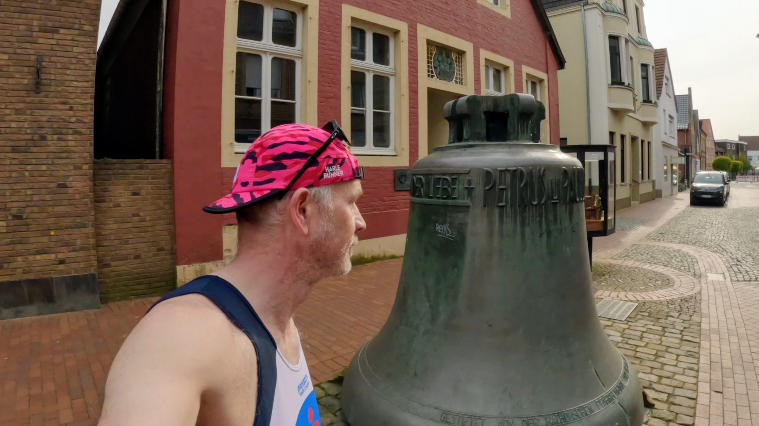 Man wearing a pink camo cap and running vest stands beside a large historic bell on a cobblestone street.