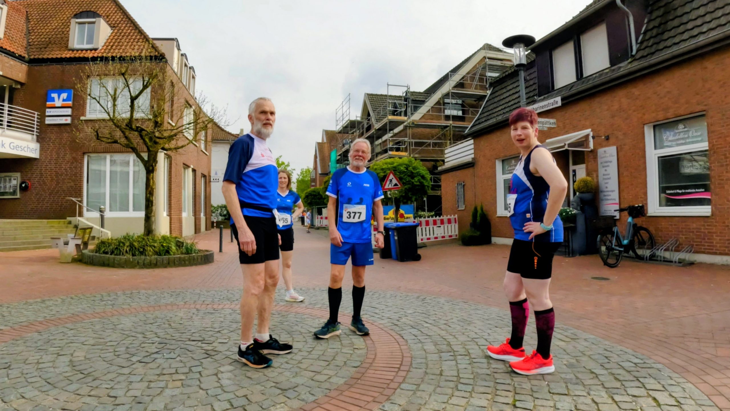 Group of four runners in blue athletic outfits standing on a circular cobblestone plaza in a town street, race bibs visible around their waists.