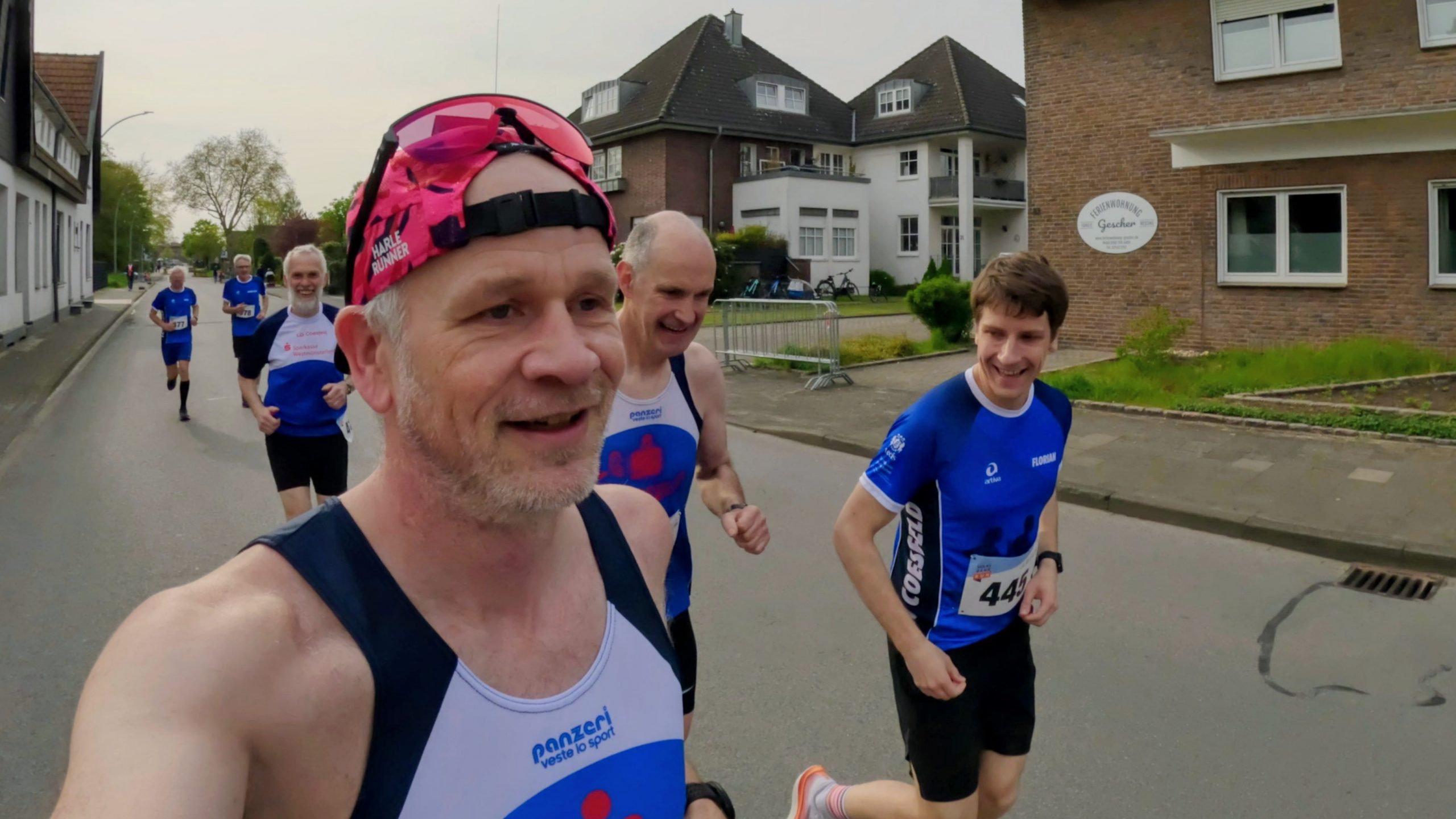 Close-up of a man in a pink cap taking a selfie while running, with a group of runners in blue jerseys behind him on a residential street downtown park area behind.