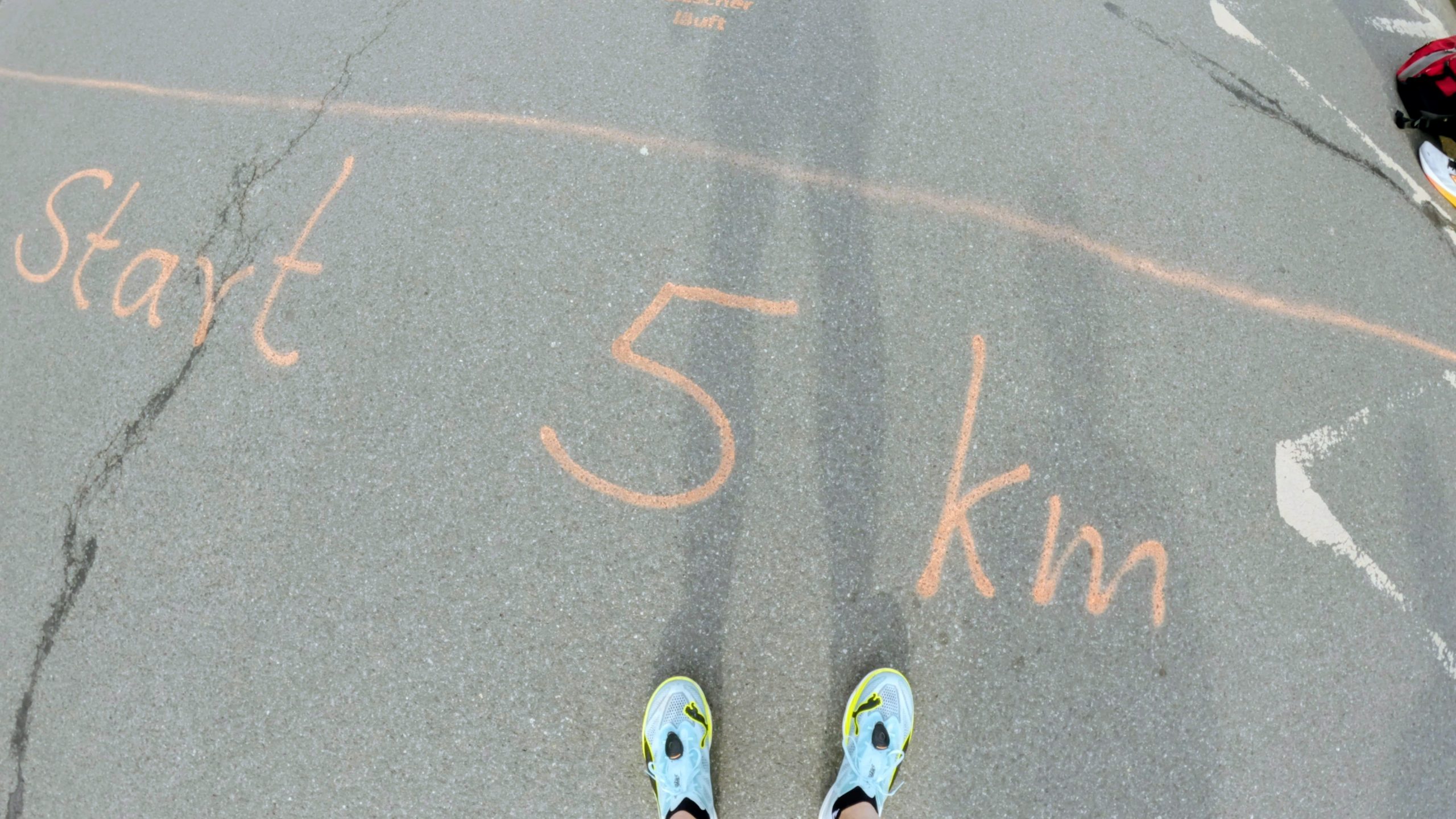 Person standing on a paved path with chalk markings reading 'Start' and '5 Km' and long shadow stretching ahead, shoes visible at the bottom as if about to begin a run.