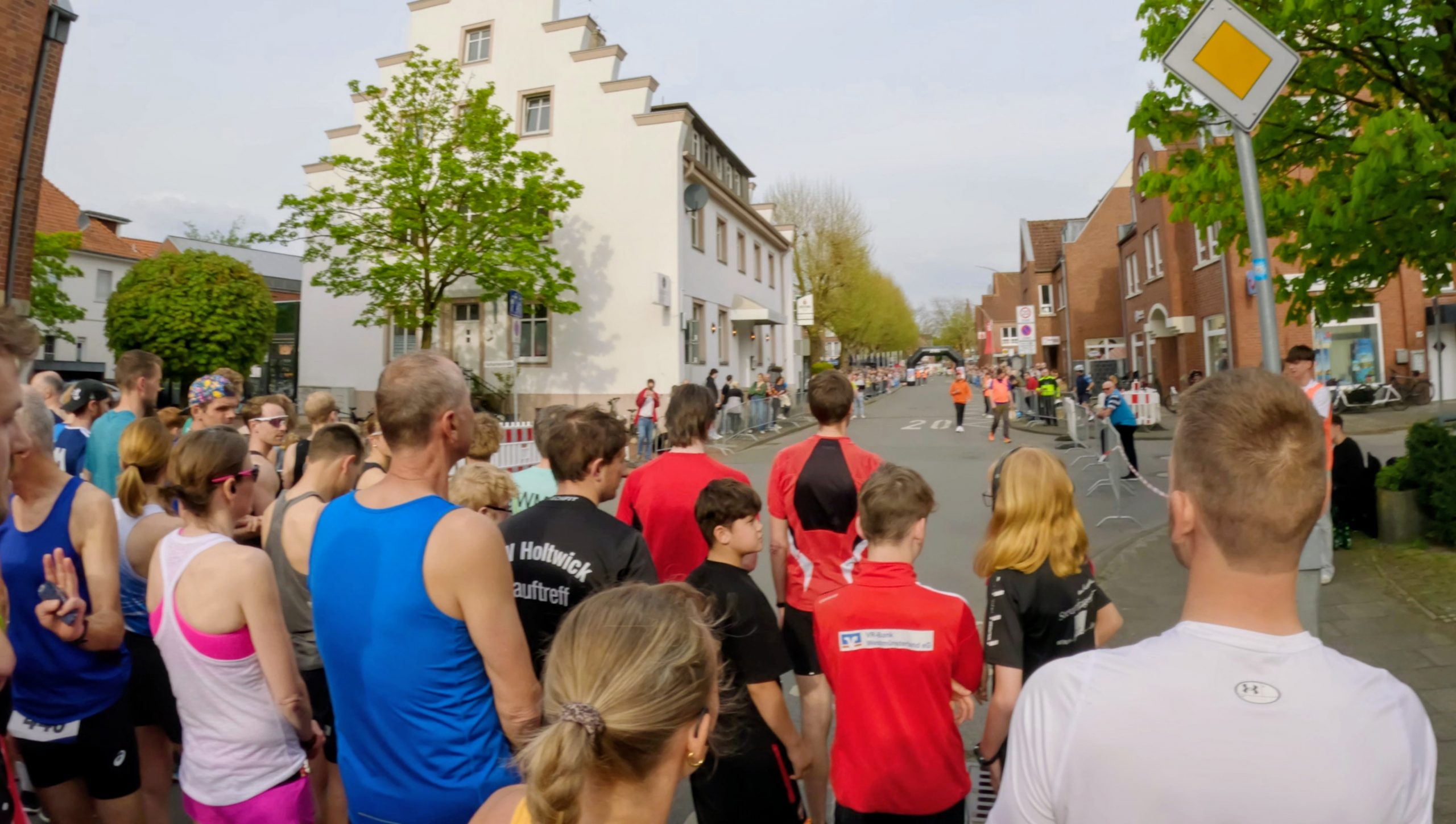 Crowd of runners and spectators along a city street, waiting at the start of a race with barriers lining the road and a finish line in the distance.