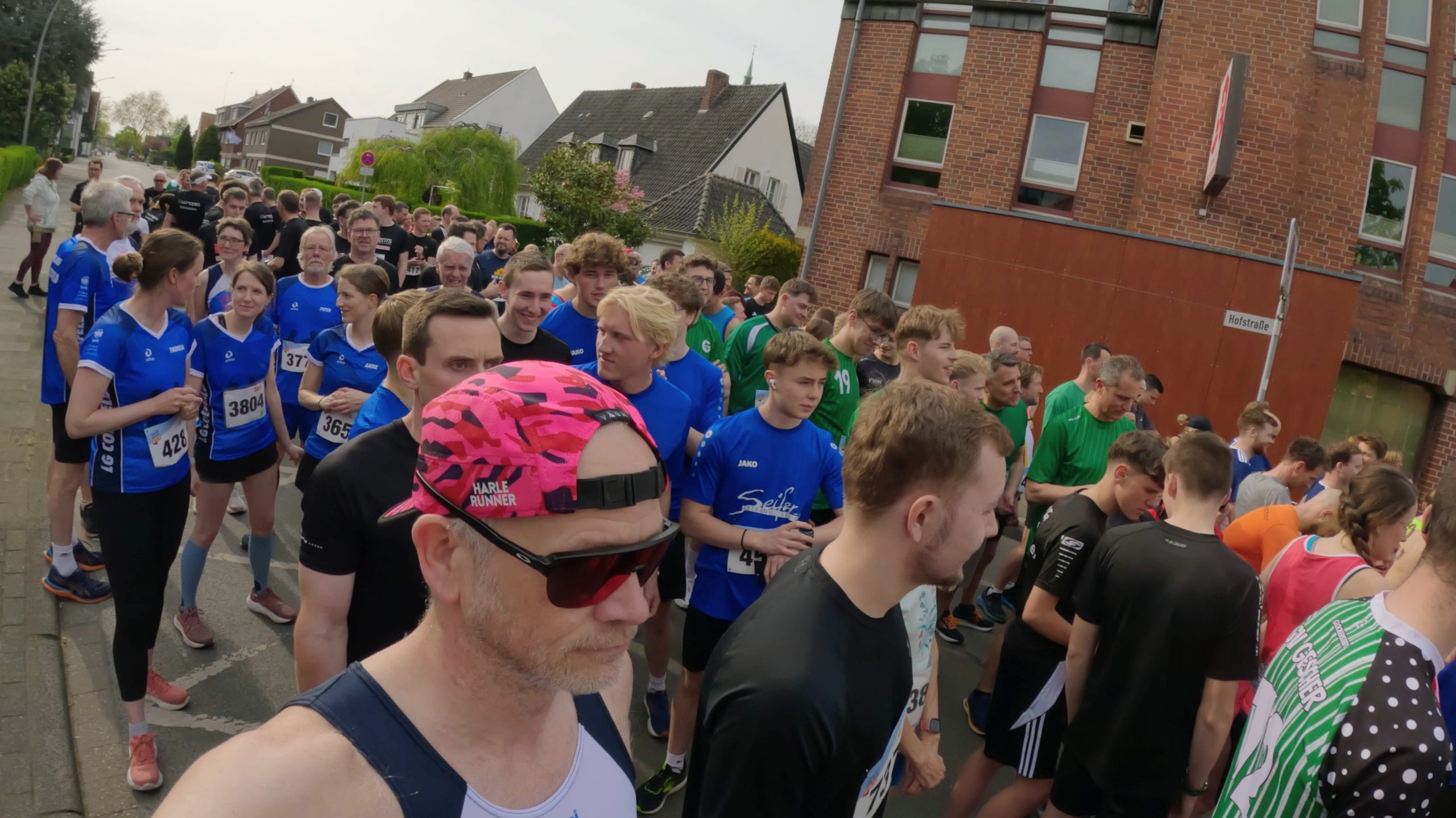 Large group of runners lined up on a street beside a brick building, wearing blue, green, and black athletic uniforms before a race start.