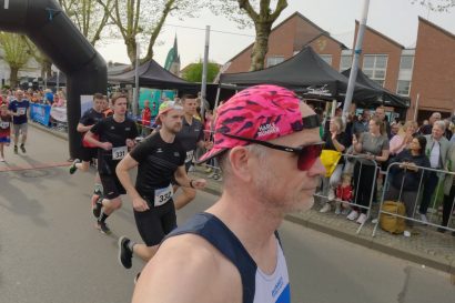 Runner with pink camouflage cap and red sunglasses leads a street race as spectators cheer behind barriers at the start area.