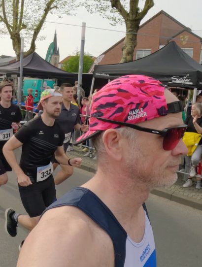 Runner with pink camouflage cap and red sunglasses leads a street race as spectators cheer behind barriers at the start area.
