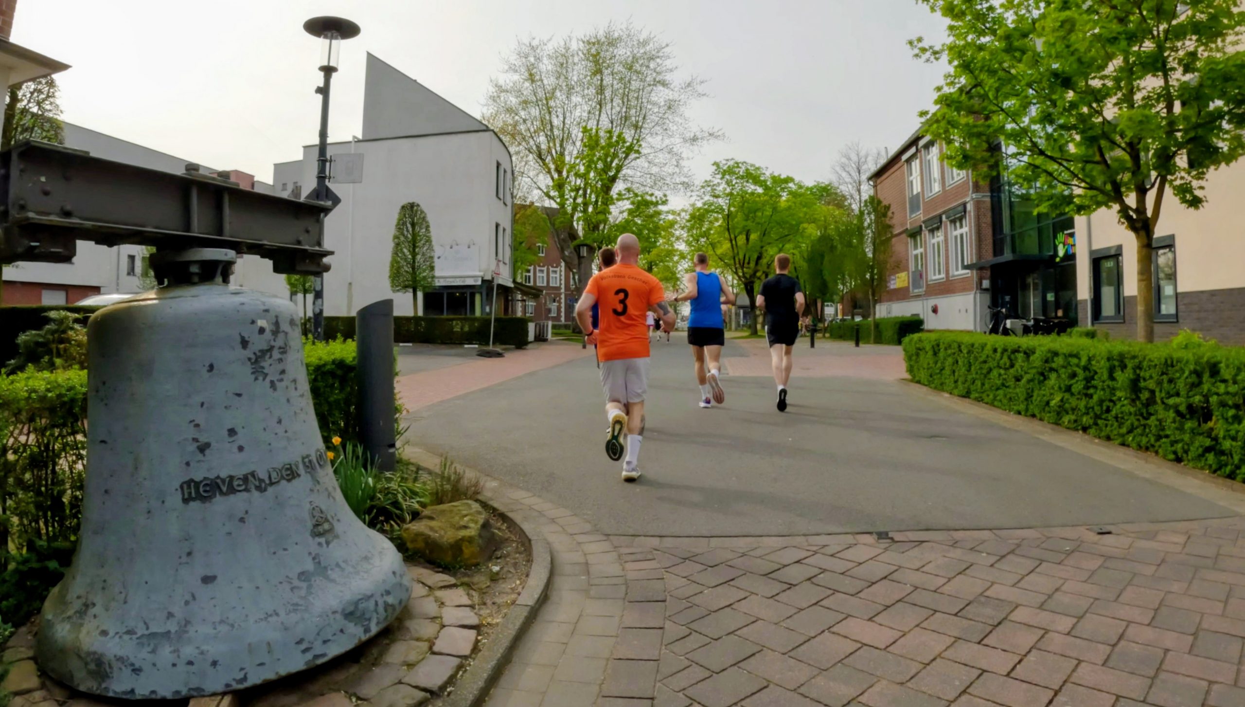 Three runners jogging along a paved street with a large bell sculpture in the foreground on the left and leafy trees lining the path in a town setting