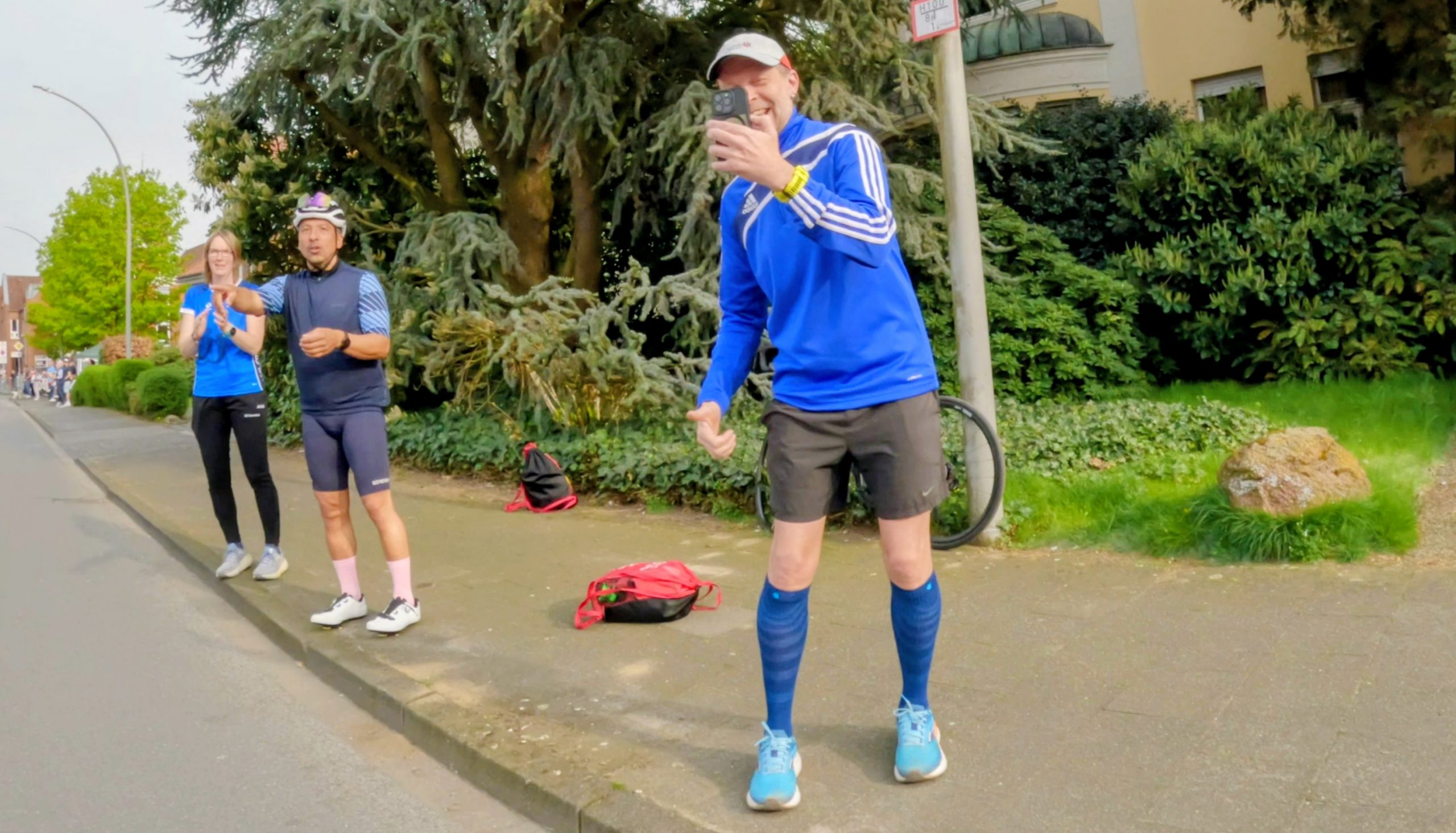 Man in blue running kit takes a selfie on a sidewalk while two teammates applaud in the background.