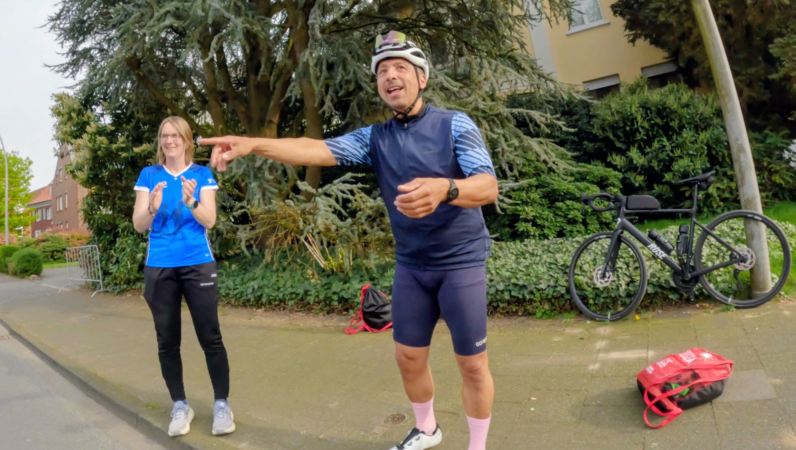 Man in cycling gear and helmet points as if giving directions, while a woman in blue claps beside him; a road bike rests against a hedge nearby.