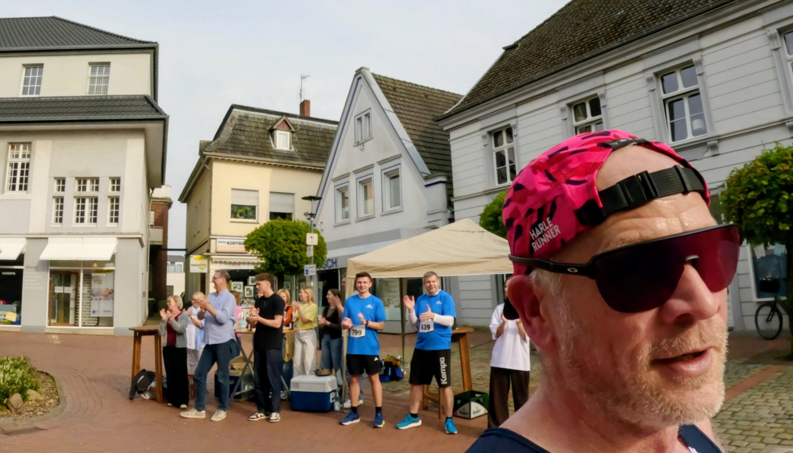 Close-up of a man in a pink cap and sunglasses in the foreground, with a crowd of runners and spectators applauding in a town square behind him.