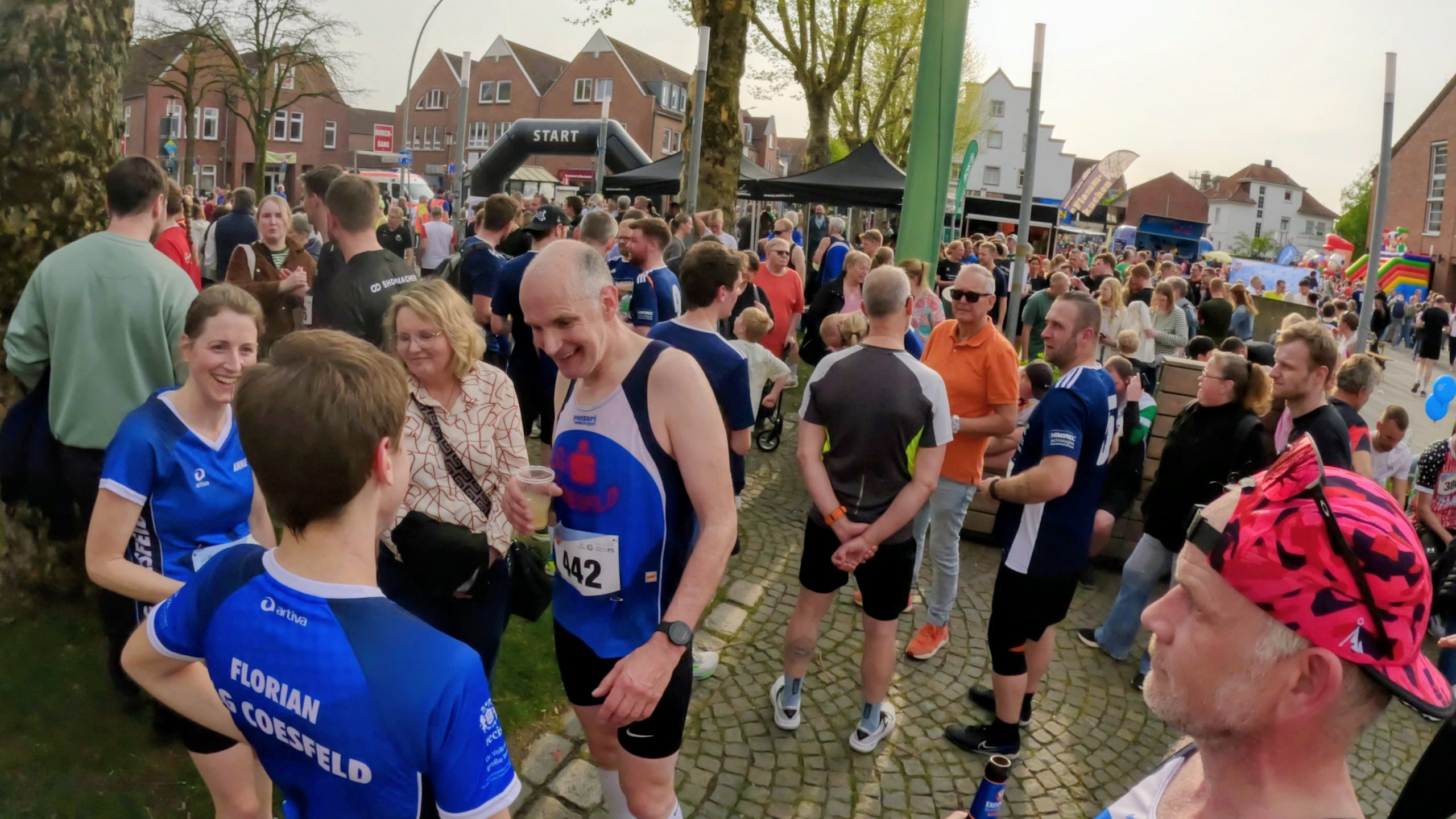 Crowd of runners and spectators at a community race start, with a black inflatable START arch in the background and people chatting on a cobblestone street.