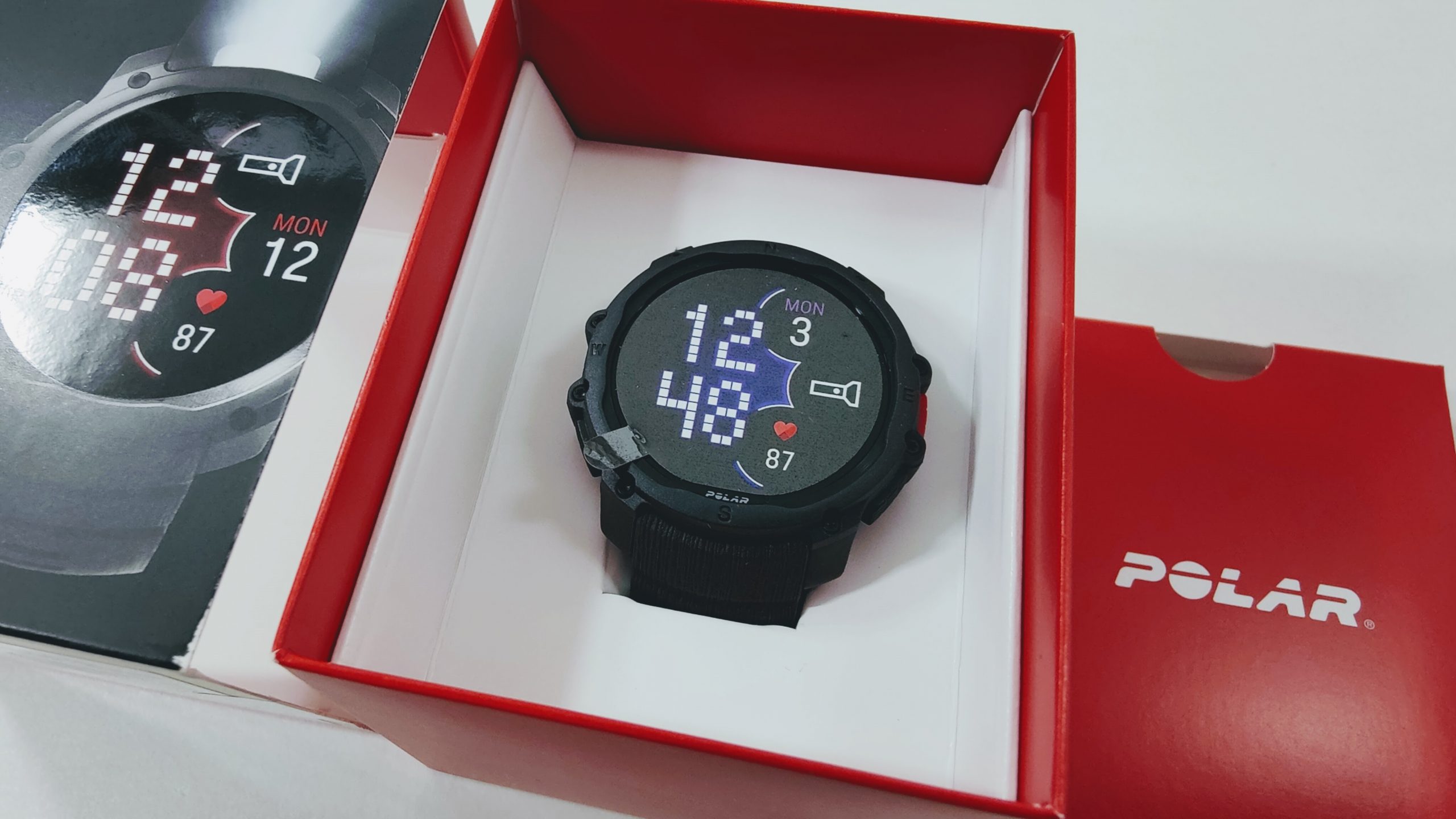 Polar smartwatch in black rugged casing inside red box packaging, display shows 12:48, Mon 3, HR 87, with heart icon and data symbols