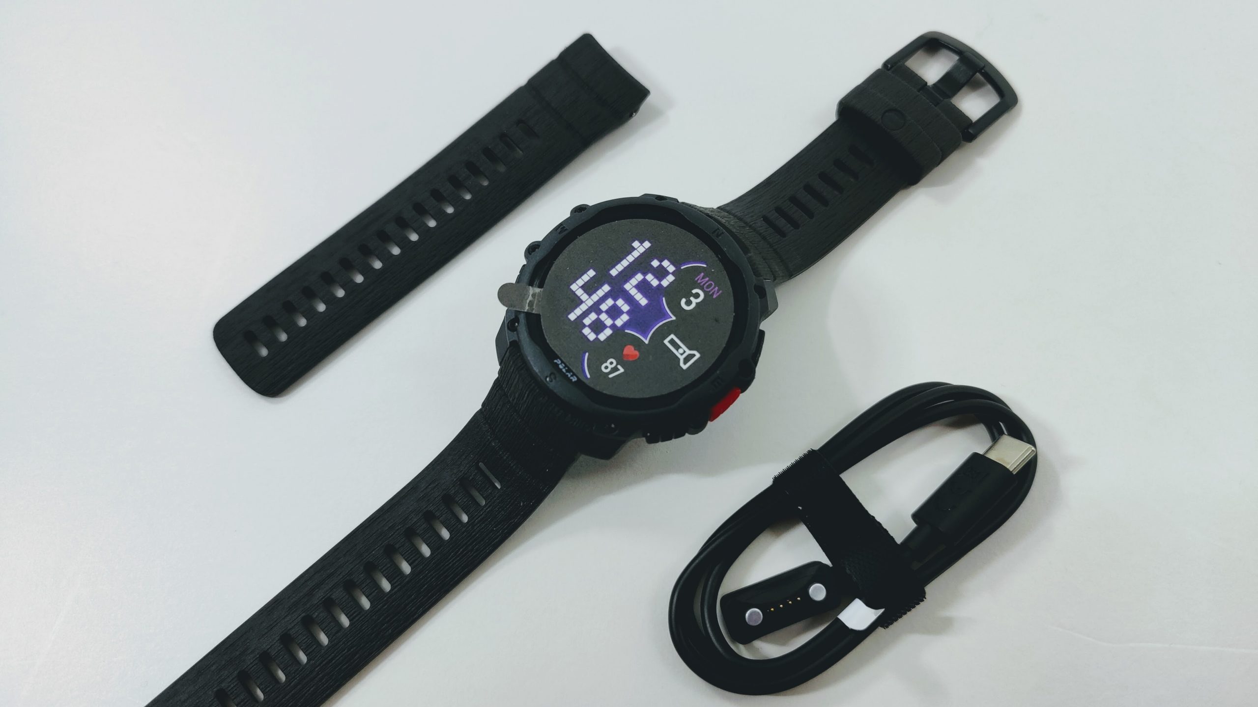 Smartwatch with a round digital display, two black replacement bands, and a USB charging cable on a light surface