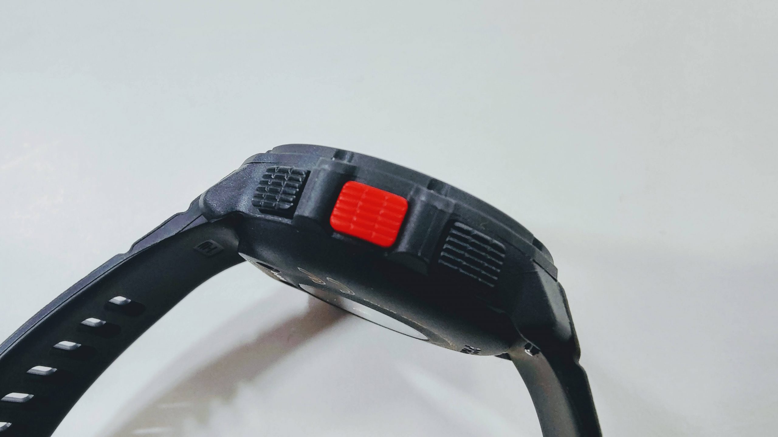 Close-up of a black sports watch from the side, showing a bright red rectangular button and textured controls on the case.