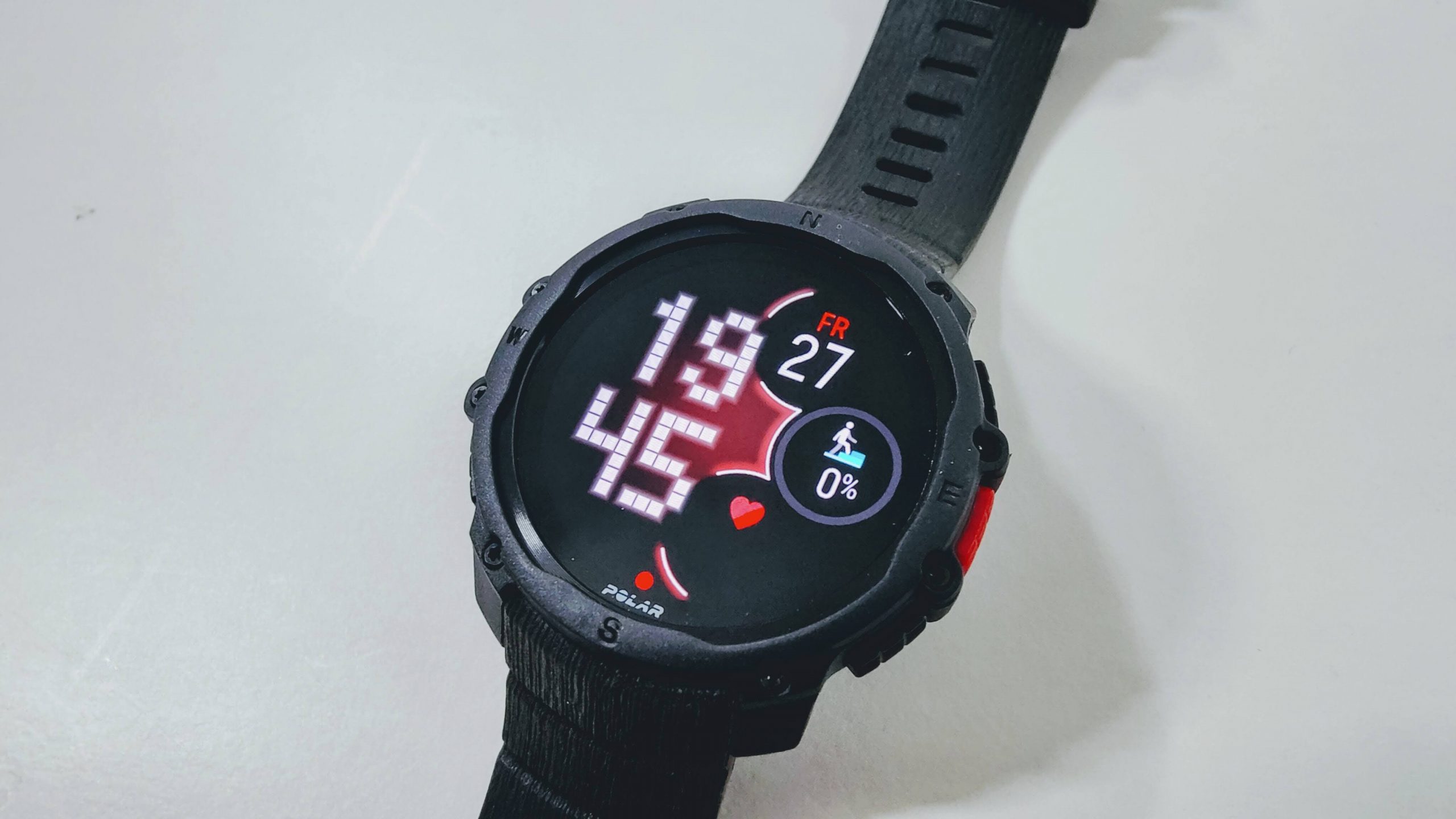 Rugged black sports smartwatch on a strap; displays time 19:45, date Fri 27, and a circular activity indicator showing 0% with a running icon and heart symbol nearby.