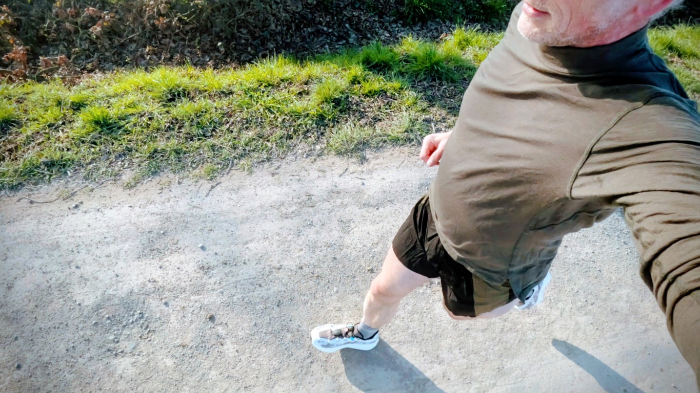 A person running on a gravel path, captured from above, wearing a green long-sleeve shirt and black shorts with white sneakers beside green grass.|Top-down view of a runner on a dirt trail with greenery nearby.|Runner in olive shirt and black shorts jogging along a sunlit dirt path, viewed from above.