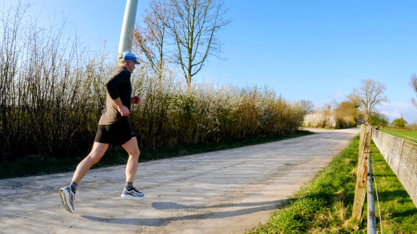 Man running along a rural dirt path on a sunny day, wearing a black shirt, shorts, and a blue cap.