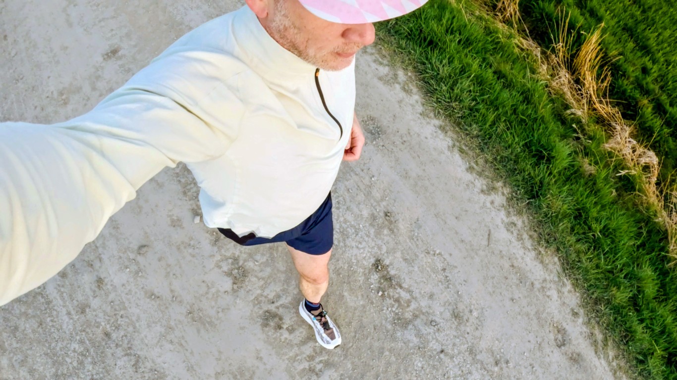 Top-down view of a person walking along a dusty trail, wearing a light jacket, pink cap, navy shorts, and white sneakers, with green grass to the side.