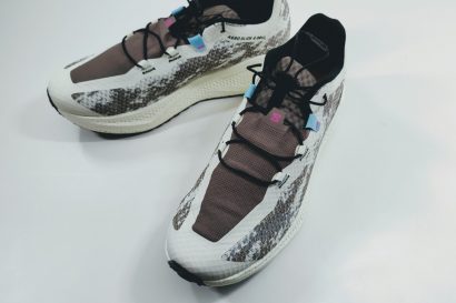 Top-down view of a pair of running shoes with white and gray patterned uppers, brown tongue areas, blue lace loops, pink logo, and black laces on a light surface.