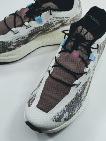 Top-down view of a pair of running shoes with white and gray patterned uppers, brown tongue areas, blue lace loops, pink logo, and black laces on a light surface.