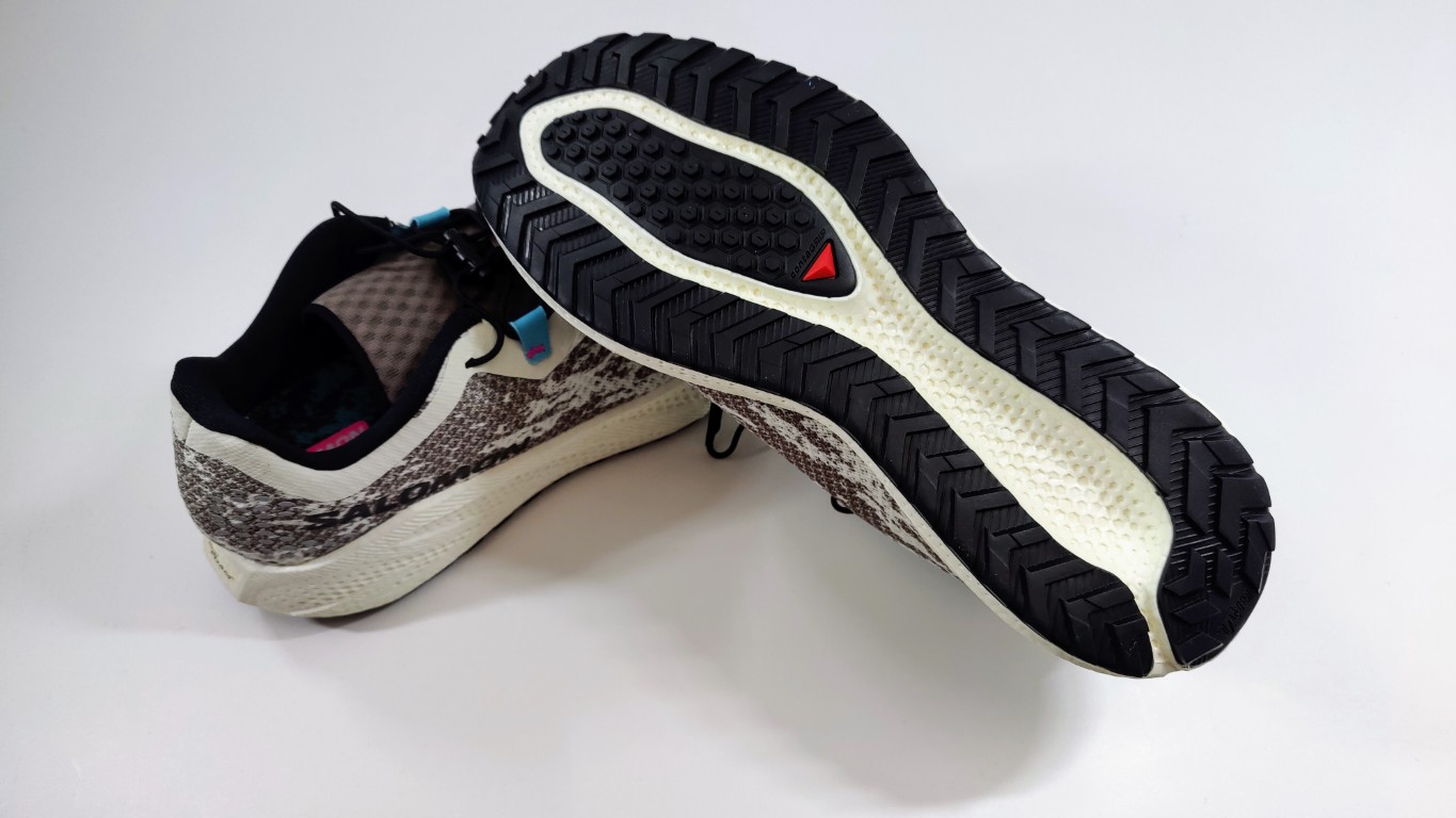 Running shoe displayed on its side showing the black outsole with deep tread and white midsole/side panel. The upper has a beige and gray knit pattern.