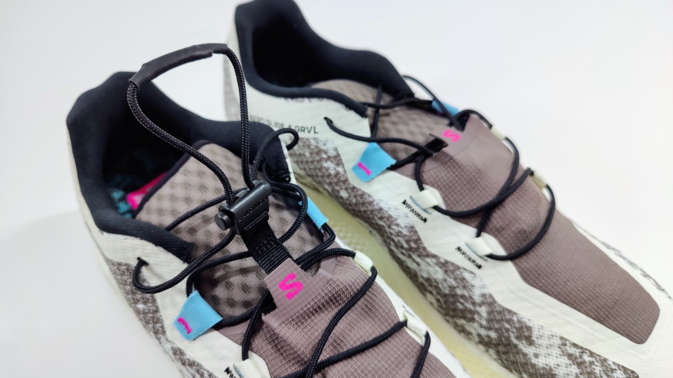 Pair of trail running shoes with taupe and beige uppers, black laces, and pink logo accents, viewed from above on a light background.