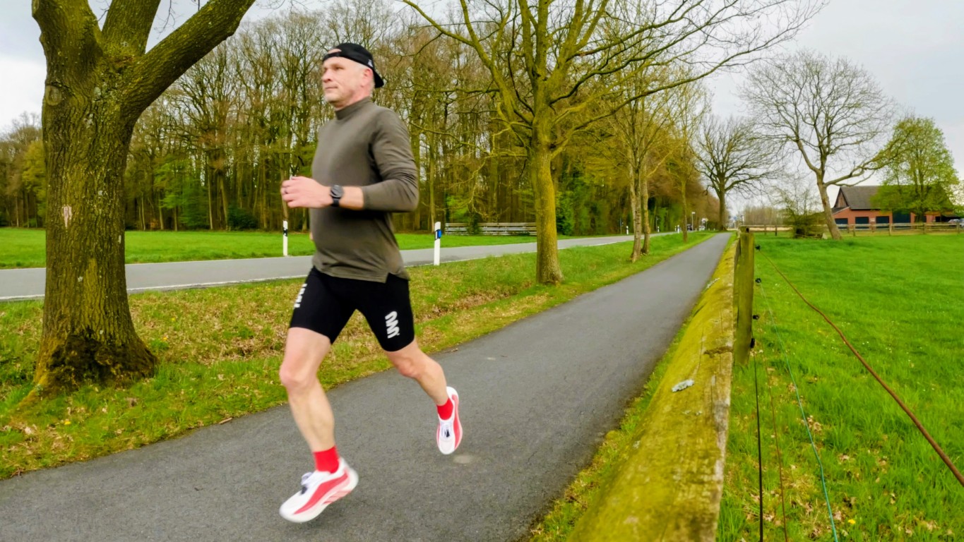 Man jogging on a paved park path, wearing an olive shirt, black shorts, red socks and white sneakers among trees. (informative)
