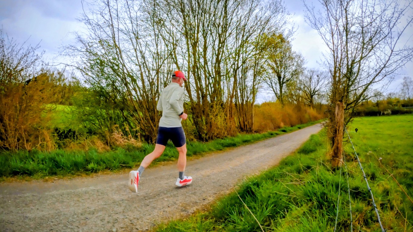 Runner in a light jacket and red cap jogging along a gravel country road with leafless trees lining the path.
