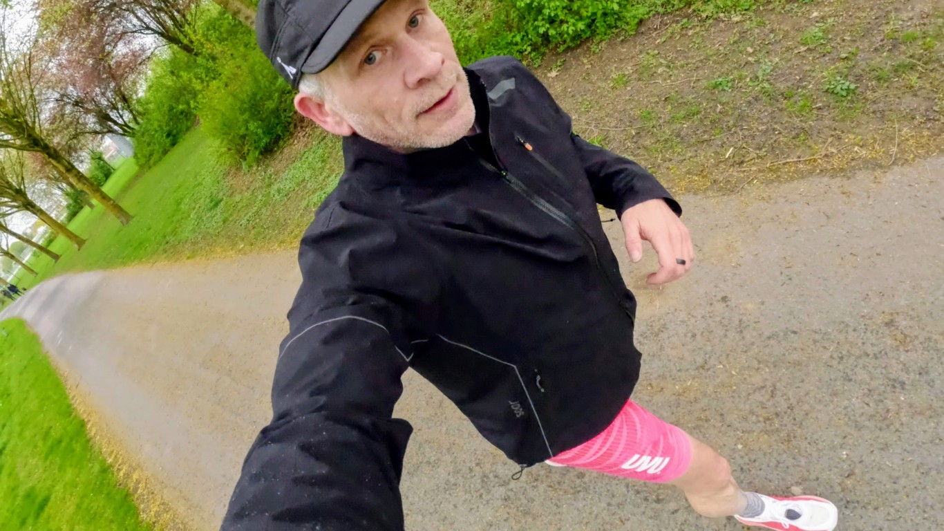 Man in a black jacket and pink shorts takes a selfie while walking/jogging on a park path with trees nearby.