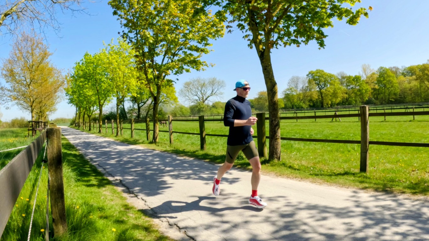 Man in athletic outfit running along a tree-lined country path on a sunny day, wearing a blue cap, black top, dark shorts, and red socks.