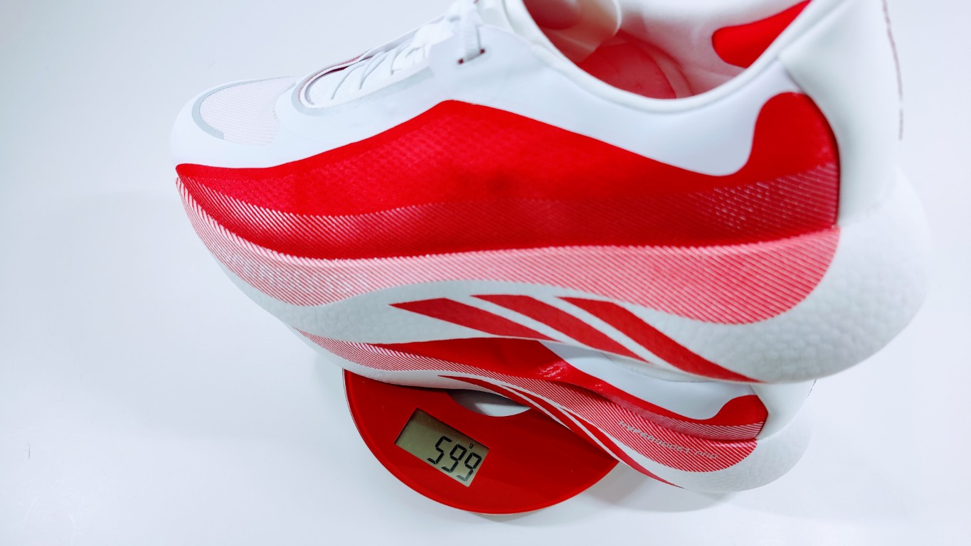 Close-up of a white and red athletic shoe on a red digital scale, the display reads 59.9 g (or 59.9).