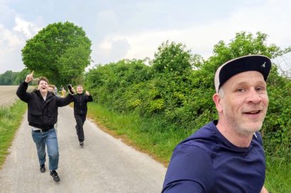 Man in a blue shirt takes a selfie on a rural road as three friends in dark jackets run toward the camera, smiling and cheering nearby trees and hedges.