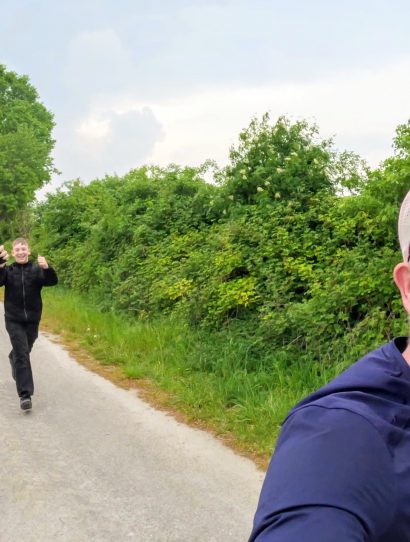 Man in a blue shirt takes a selfie on a rural road as three friends in dark jackets run toward the camera, smiling and cheering nearby trees and hedges.