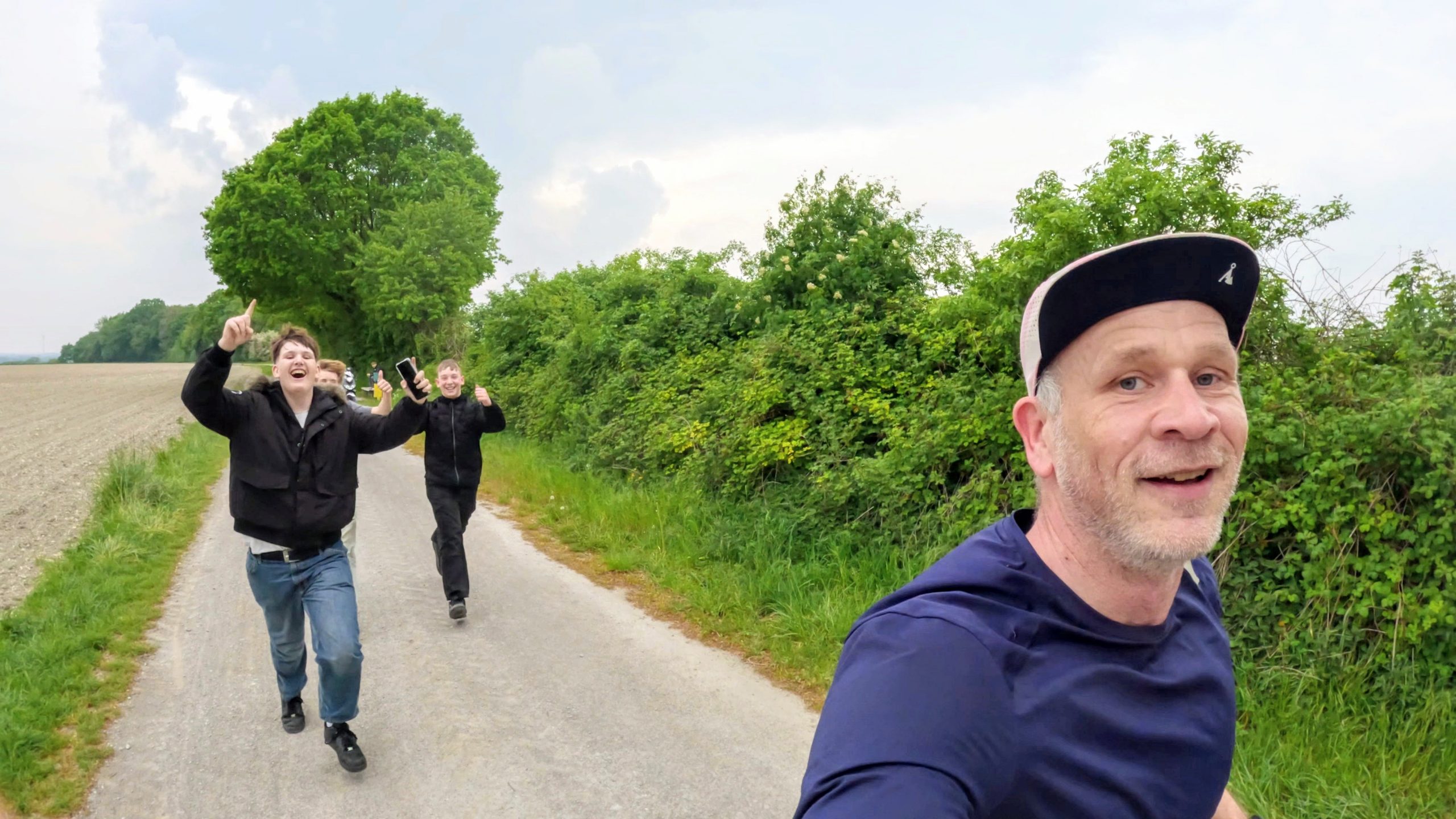 Man in a blue shirt takes a selfie on a rural road as three friends in dark jackets run toward the camera, smiling and cheering nearby trees and hedges.