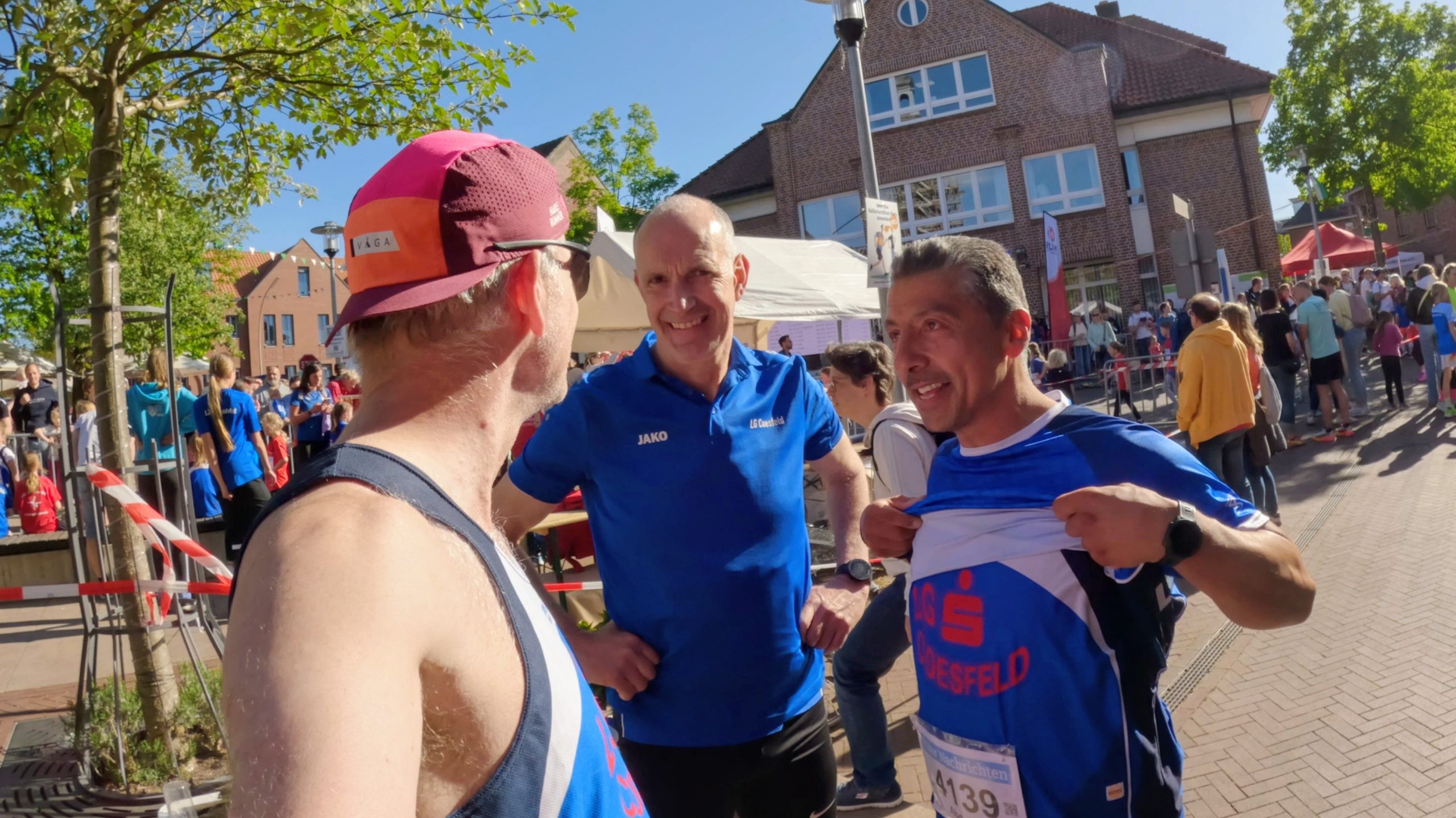 Two runners in blue jerseys chat with a man wearing a burgundy cap at a sunny street race, with spectators and tents in the background.