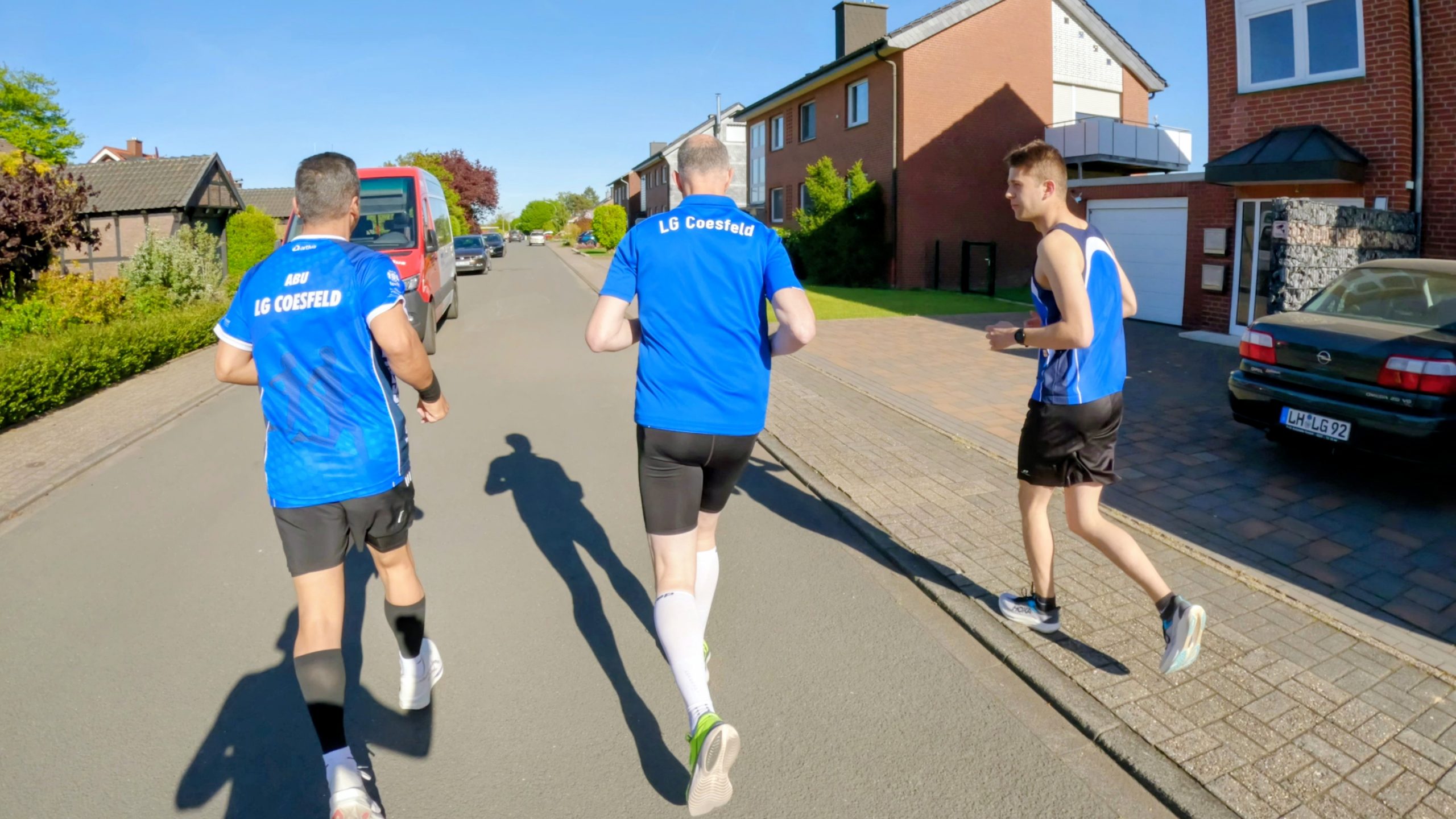 Three runners in matching blue jerseys sprint down a sunny residential street, seen from behind with houses and parked cars along the curb.