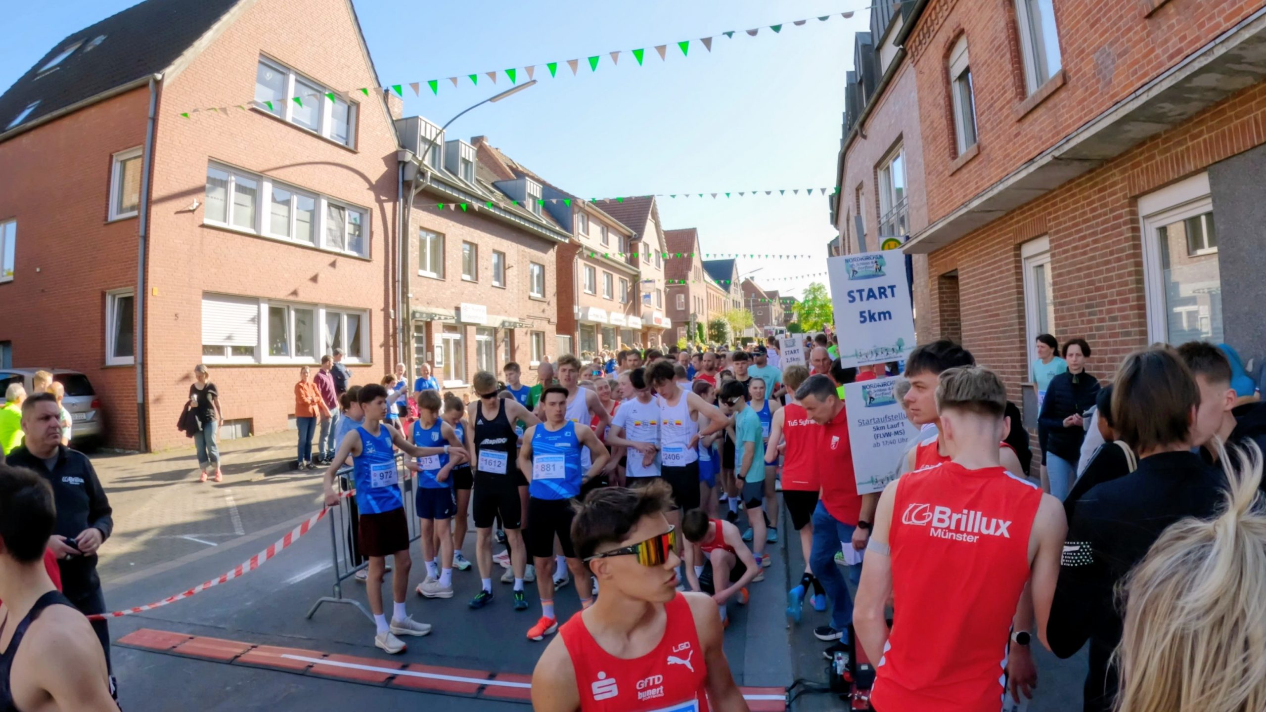 Crowded runners at the street start of a 5km race in a residential area, brick buildings and festive bunting overhead.