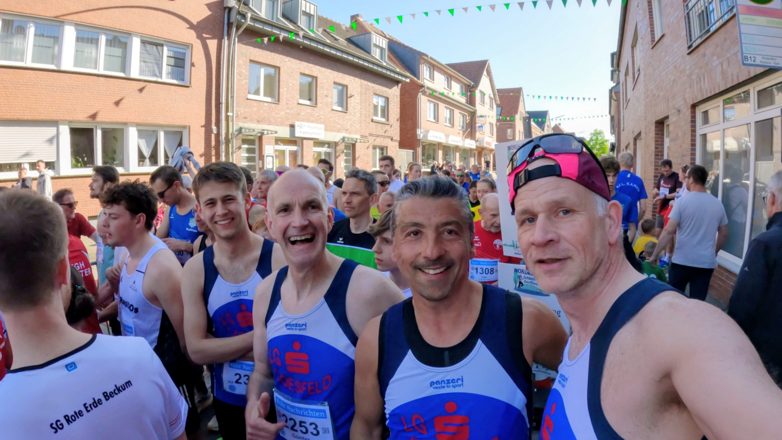 Runners in a street race line up on a sunny brick street, with spectators and green bunting overhead.