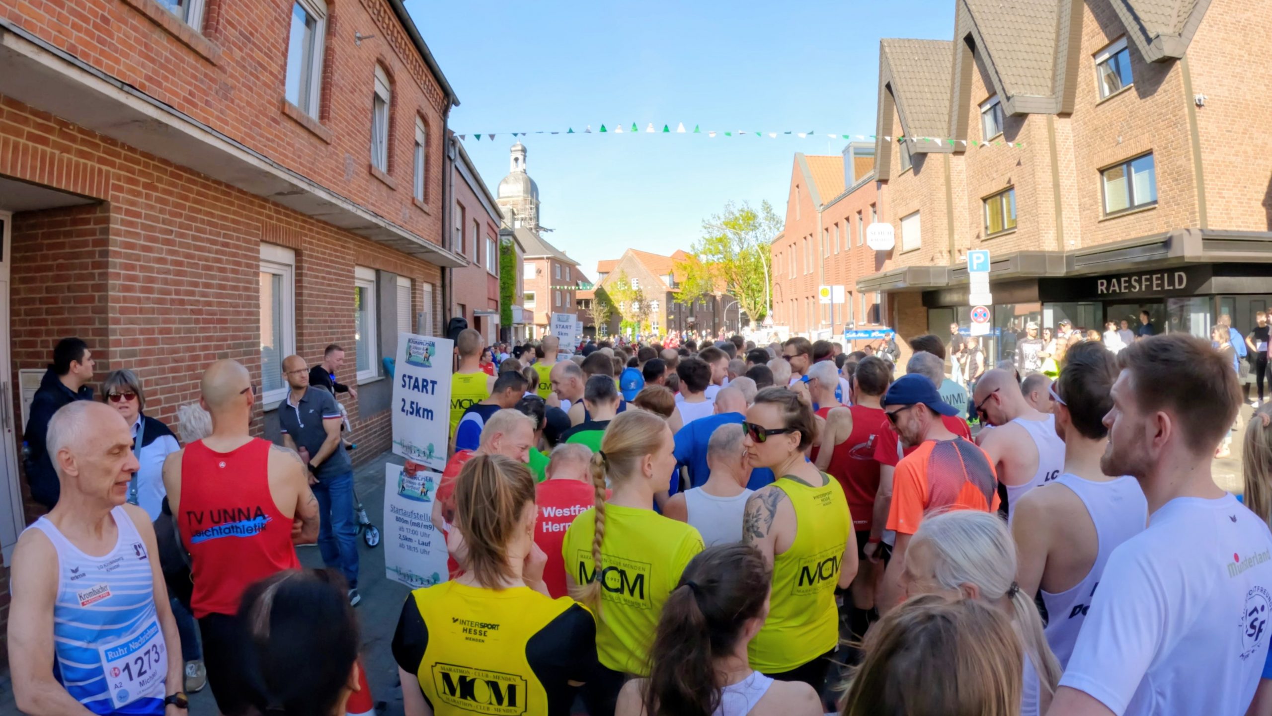 Crowd of runners at a city street start, with brick buildings on both sides and spectators watching.