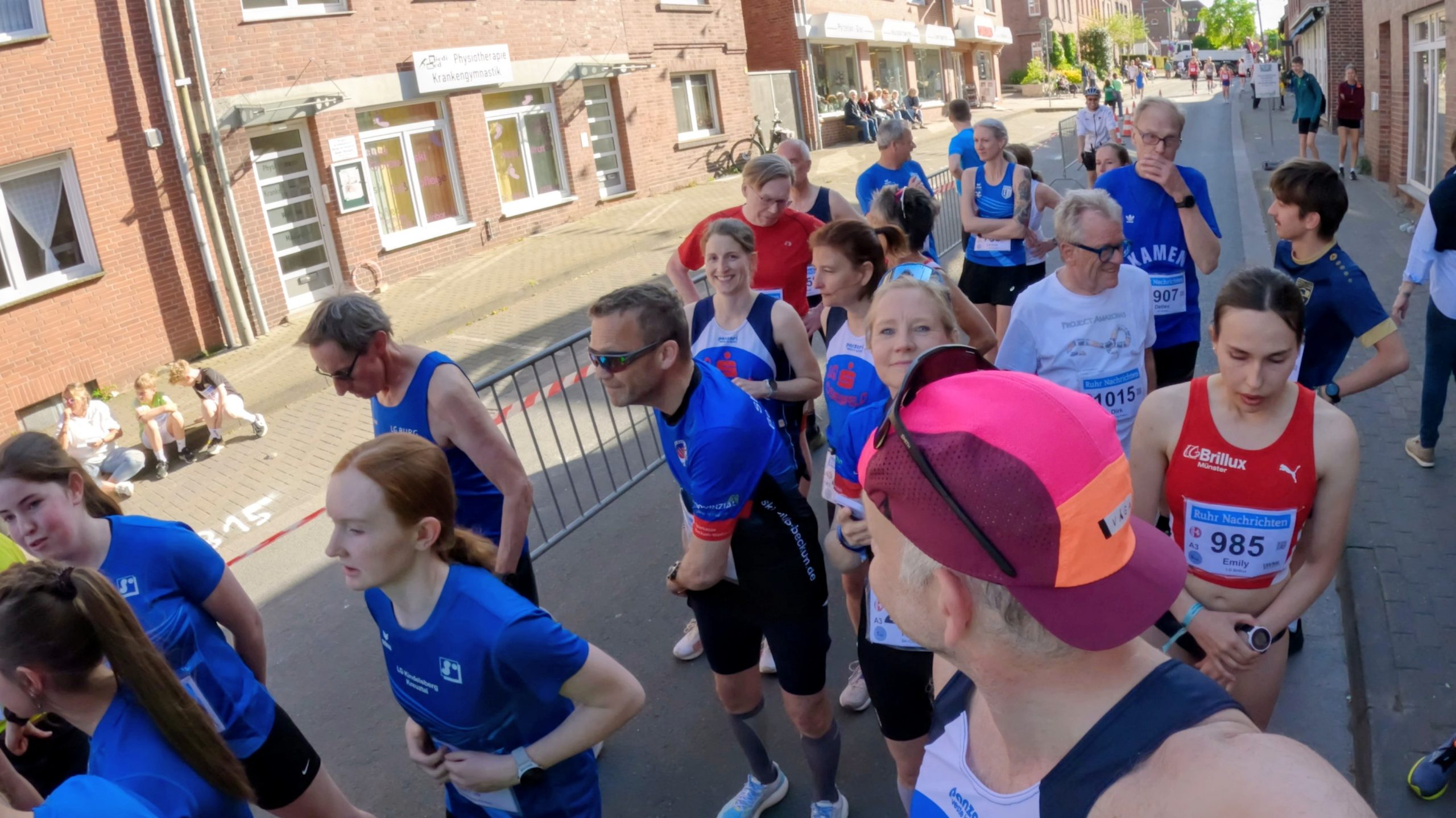 Pack of runners in a street race moving past brick storefronts on a sunny day, with spectators sitting along the curb in the background.