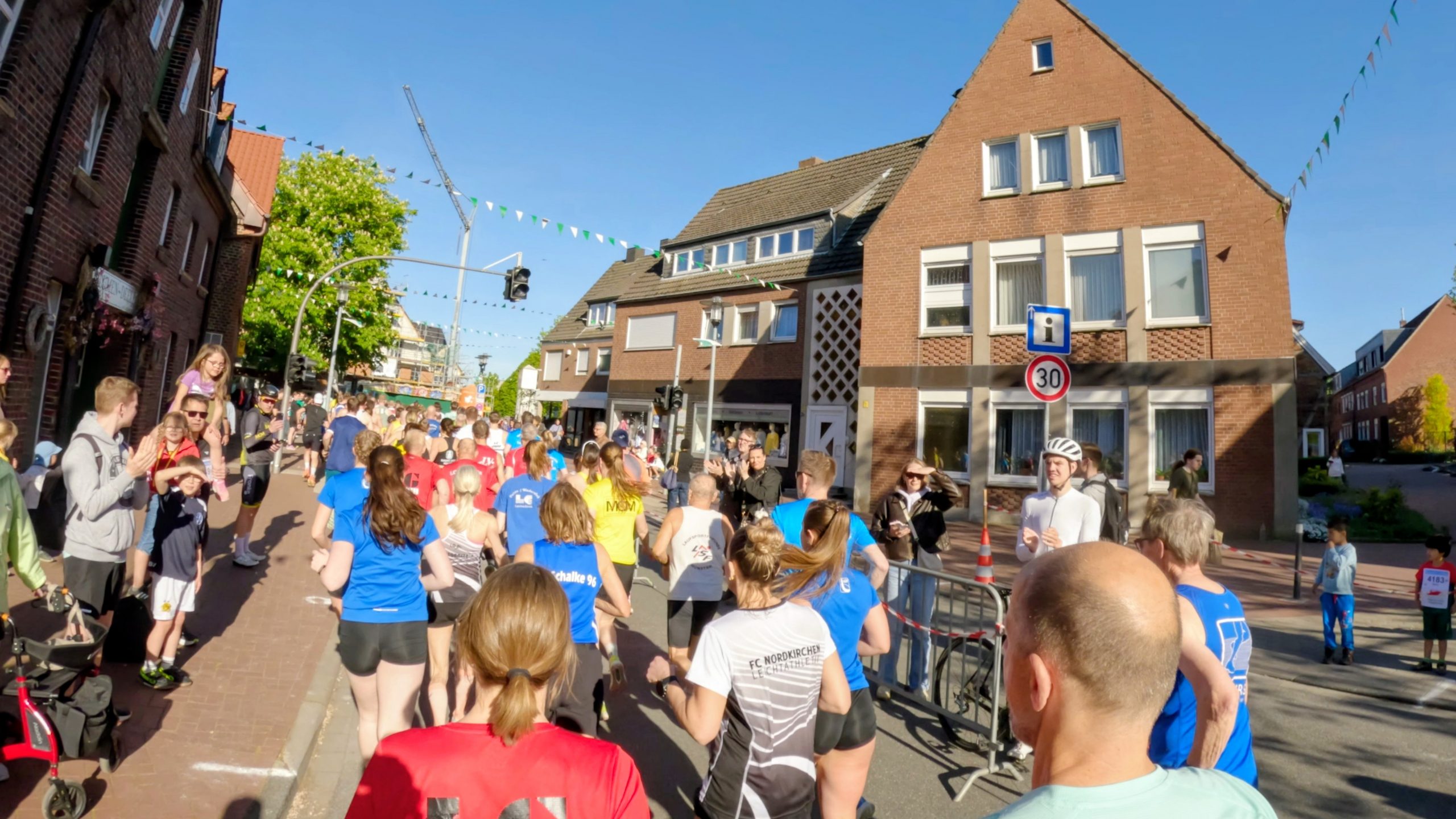 Crowd of runners in a sunny street race passing brick houses with festive bunting overhead.