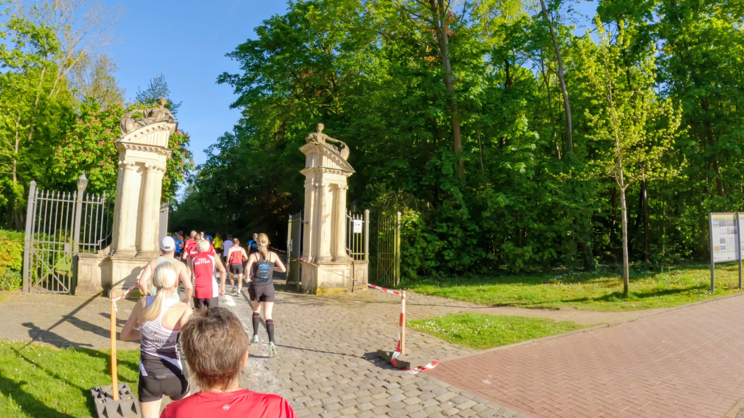 Runners in a race enter a park through tall decorative stone gates on a sunny day, with green trees and a cobblestone path easing into the forested area.