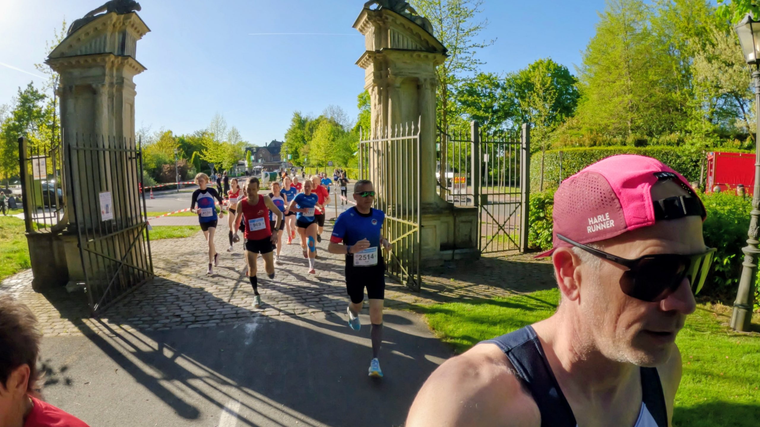 Runners in a race exit through an ornate stone gate with iron fencing on a sunny day; bib numbers visible on their shirts; photographer in foreground wearing a pink cap.