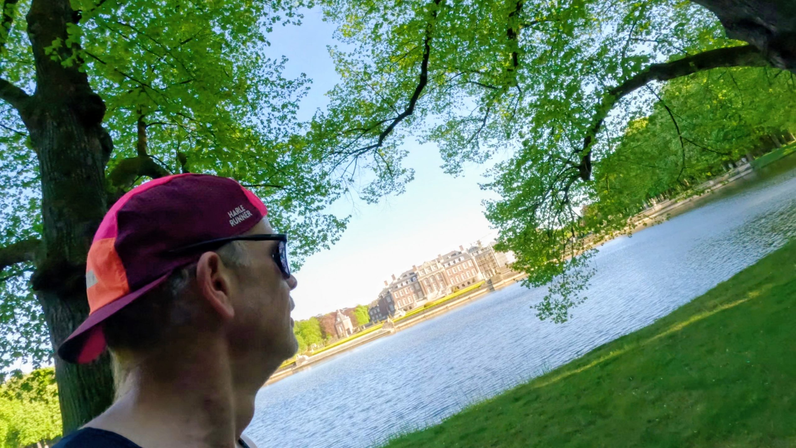 Side profile of a person wearing a pink running cap and sunglasses by a calm lake, with green trees and buildings in the background.