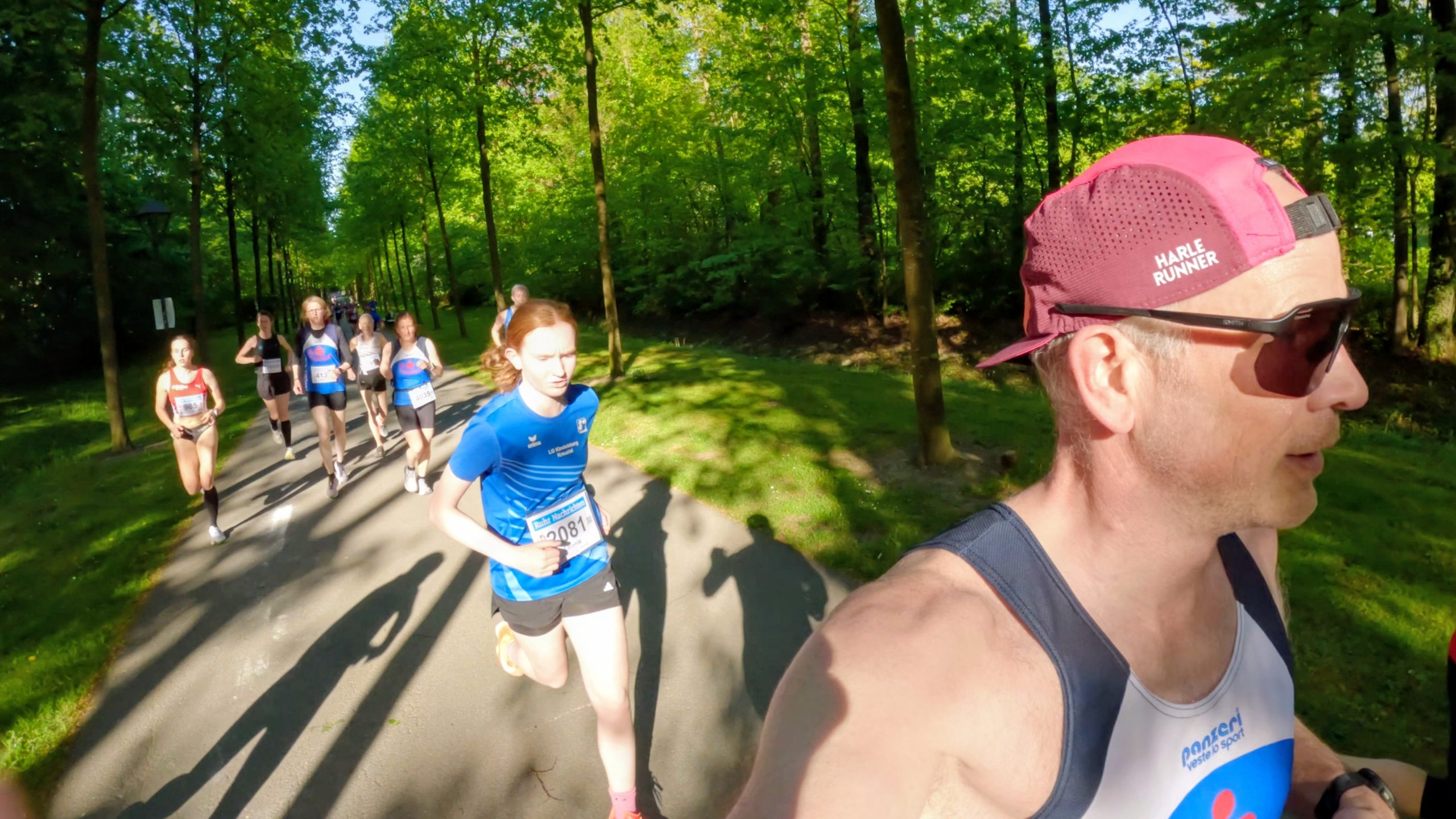 Lead runner in a pink cap and sunglasses on a tree-lined park road, with a group of runners behind him in a race.