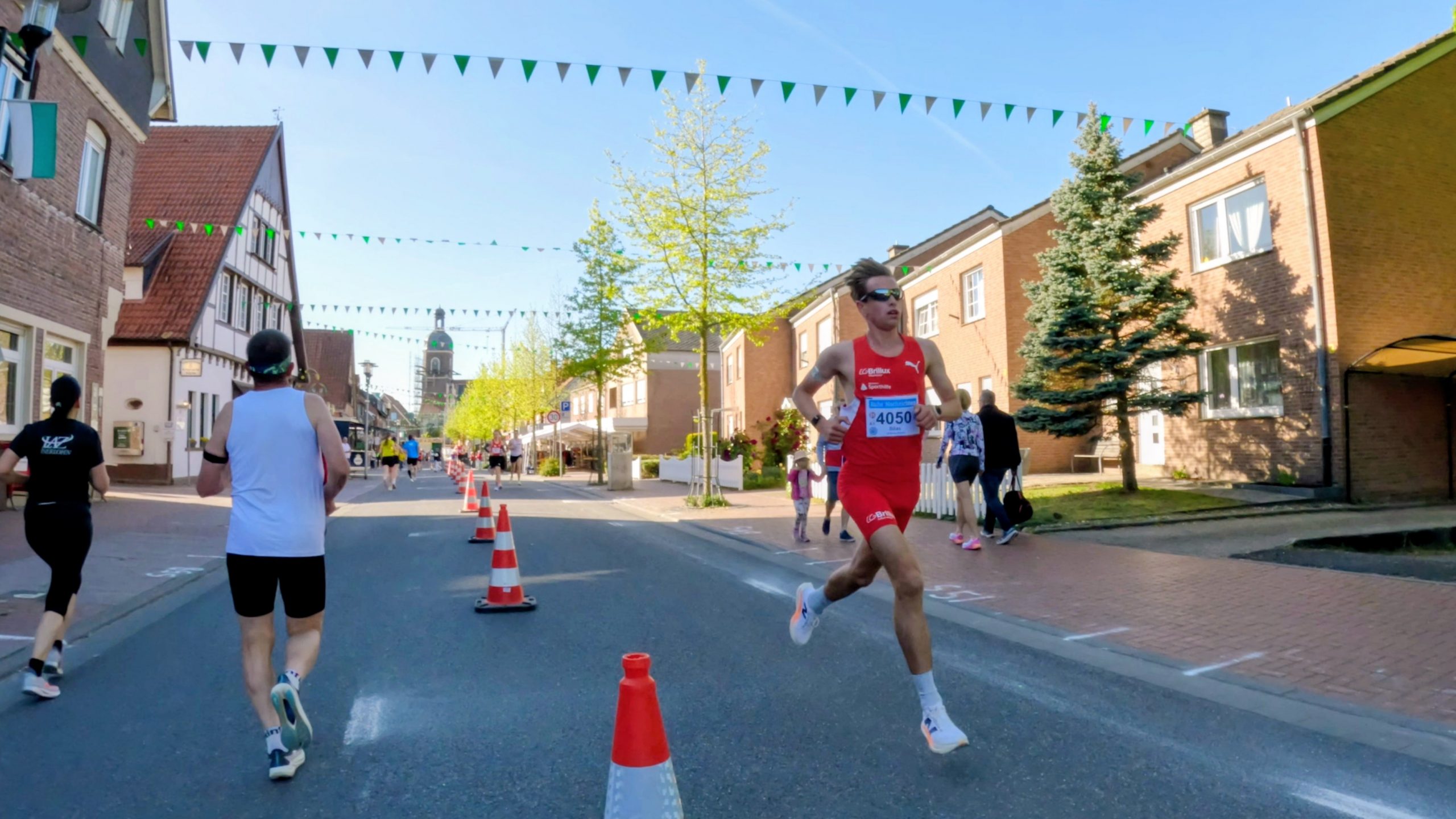 Male runner in red kit with bib 4050 leads a street race past orange traffic cones on a sunny town street.