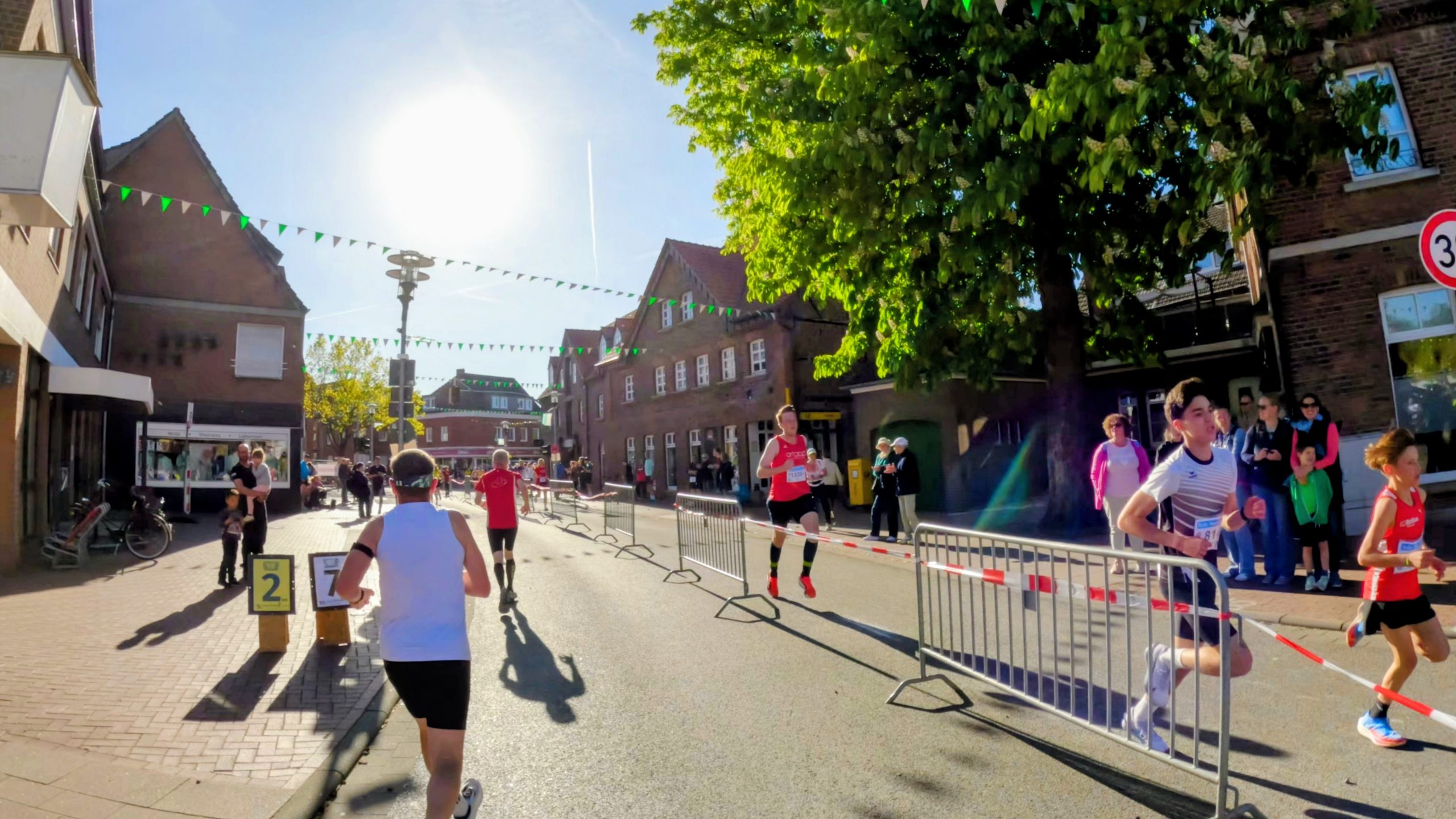Runners race down a sunny town street lined with brick buildings, barriers and cheering spectators on the sidewalk.