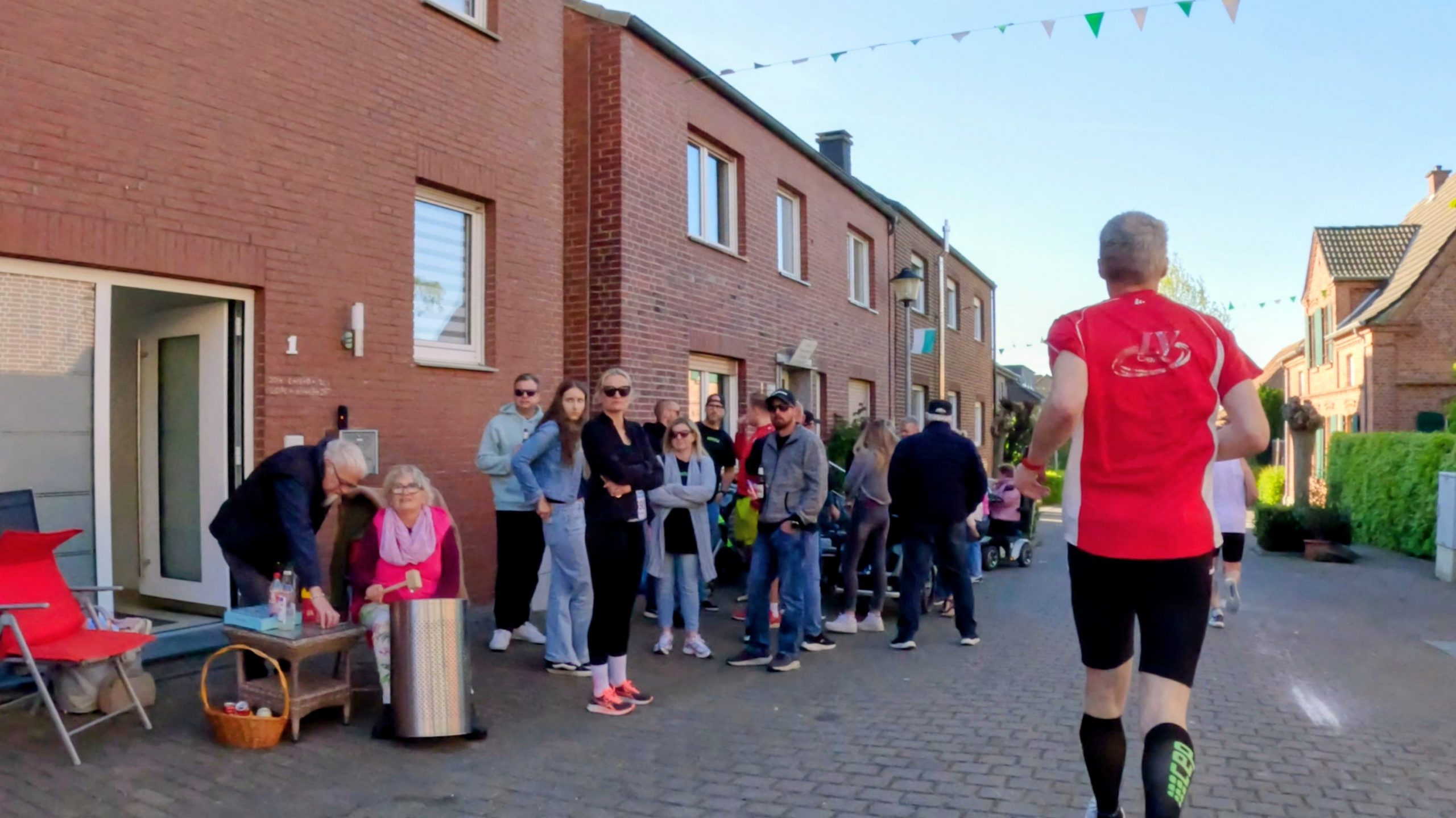 Runner in a red shirt seen from behind, approaching a line of spectators on a brick residential street with bunting overhead.