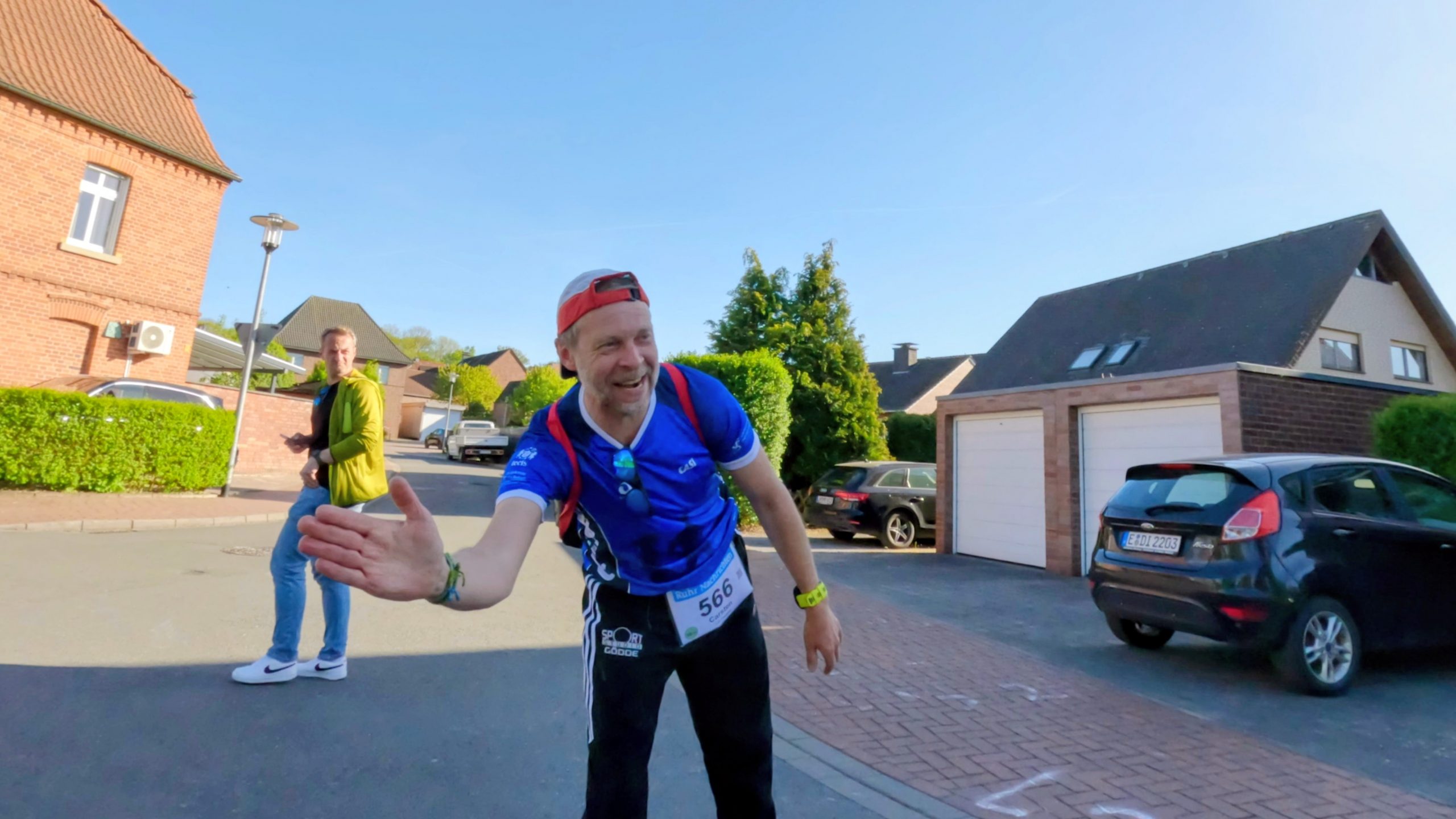 Runner in a blue cycling top extends hand toward the camera while smiling, on a suburban street during a race; spectator in yellow jacket behind him; houses and parked cars ahead.
